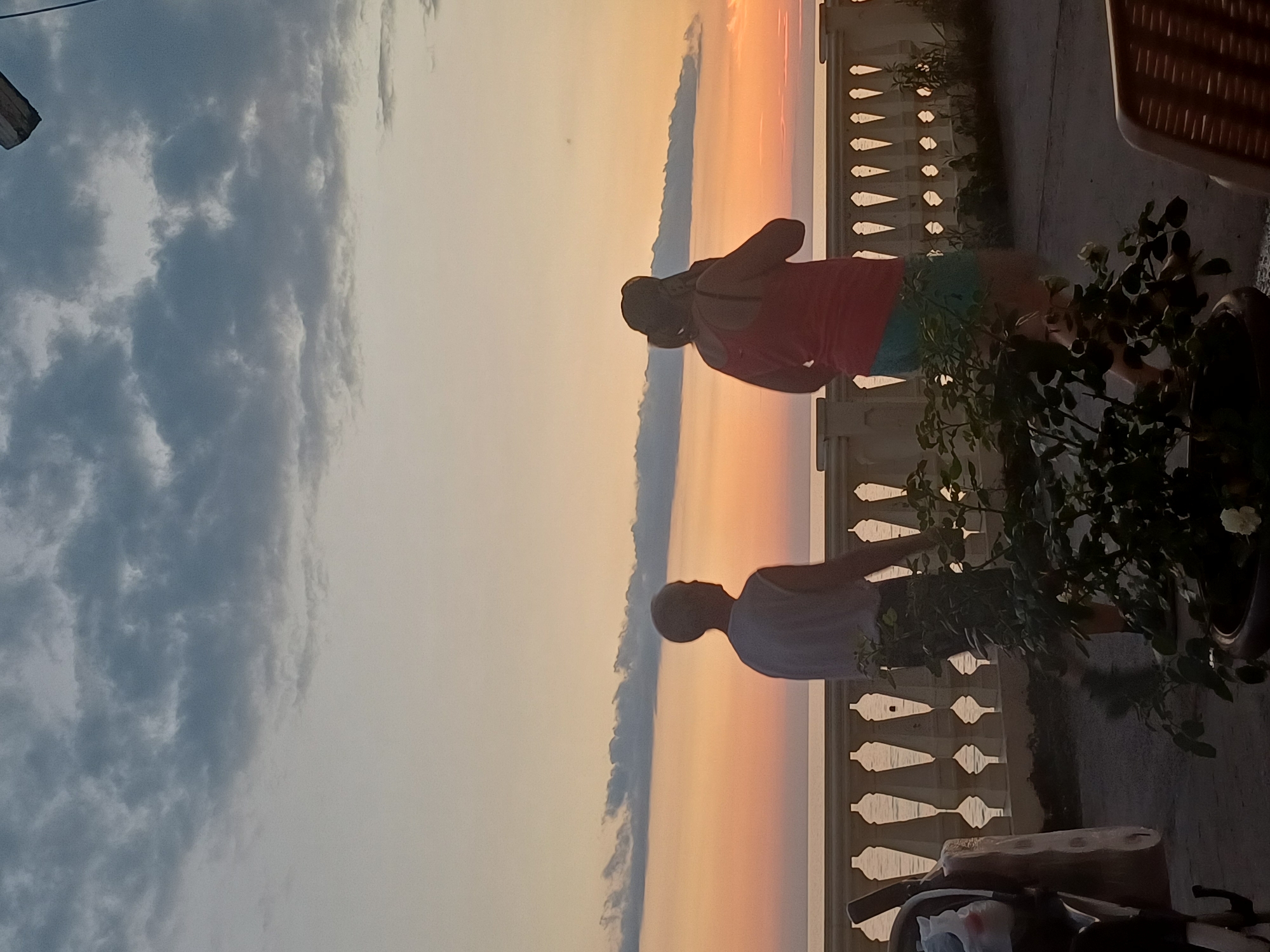 The image depicts a sunset scene on the embankment in Sochi, Russia. Two individuals are standing on the embankment, facing away from the camera, looking out at the sea. The sky is filled with dramatic clouds, with hues of orange and pink from the setting sun blending into the lighter sky above. In the foreground, a decorative white railing runs along the embankment. There are also plants in pots and some outdoor furniture visible. The overall atmosphere is tranquil and serene, typical of an evening on the Black Sea coast.