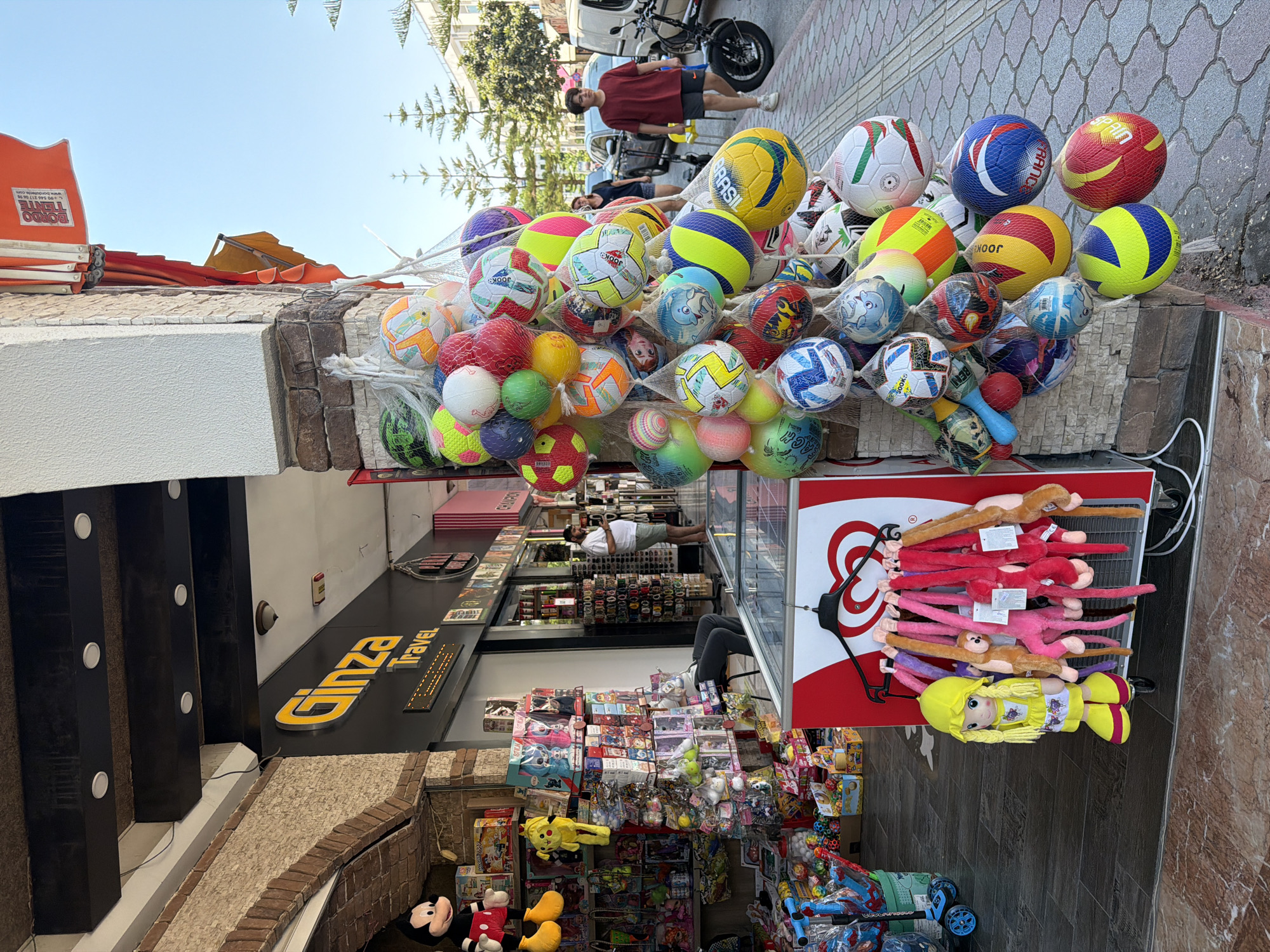 The image depicts a street scene in Kemer, Turkey, featuring a storefront with an abundance of merchandise displayed outside. Directly in front of a building with a "Ginza Travel" sign, a variety of balls, predominantly soccer and volleyballs of different sizes and colors, are hanging in netting. To the left, shelves are stocked with toys and trinkets, including a large Mickey Mouse figure and several plush toys. A yellow doll with a matching hat and shoes stands near a red and white cooler. In the background, people are walking on the sidewalk, and vehicles, including a motorcycle and cars, are visible. The environment appears to be a commercial area with a clear blue sky overhead.