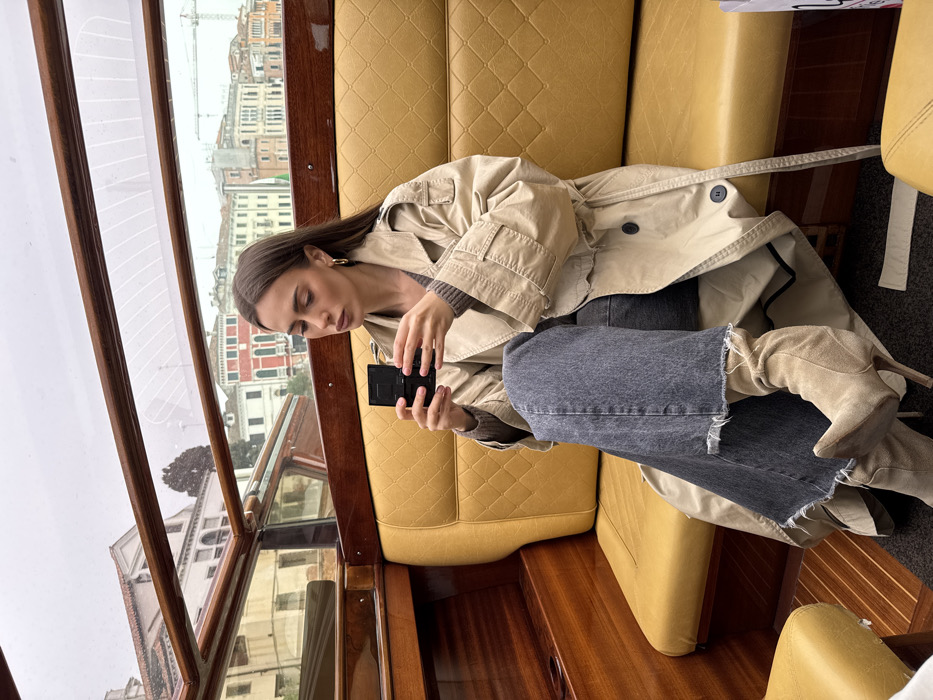 An adult female is seated within the cabin of a watercraft on a canal in Venice, Italy. She has long, dark brown hair and is wearing gold hoop earrings. Her attire includes a light beige trench coat over a dark grey knit top, dark grey wide-leg jeans with frayed hems, and light beige pointed-toe, high-heeled ankle boots. She holds a small, black rectangular compact object with both hands, directing her gaze towards its surface. The boat's interior features polished dark wood paneling, yellow diamond-quilted vinyl seating, and a clear glass canopy providing an external view. Outside, multi-story buildings with varied architectural styles and colors line the canal under an overcast sky. The white arched structure of the Ponte della Costituzione (Calatrava Bridge) is visible in the distant background, confirming the location.