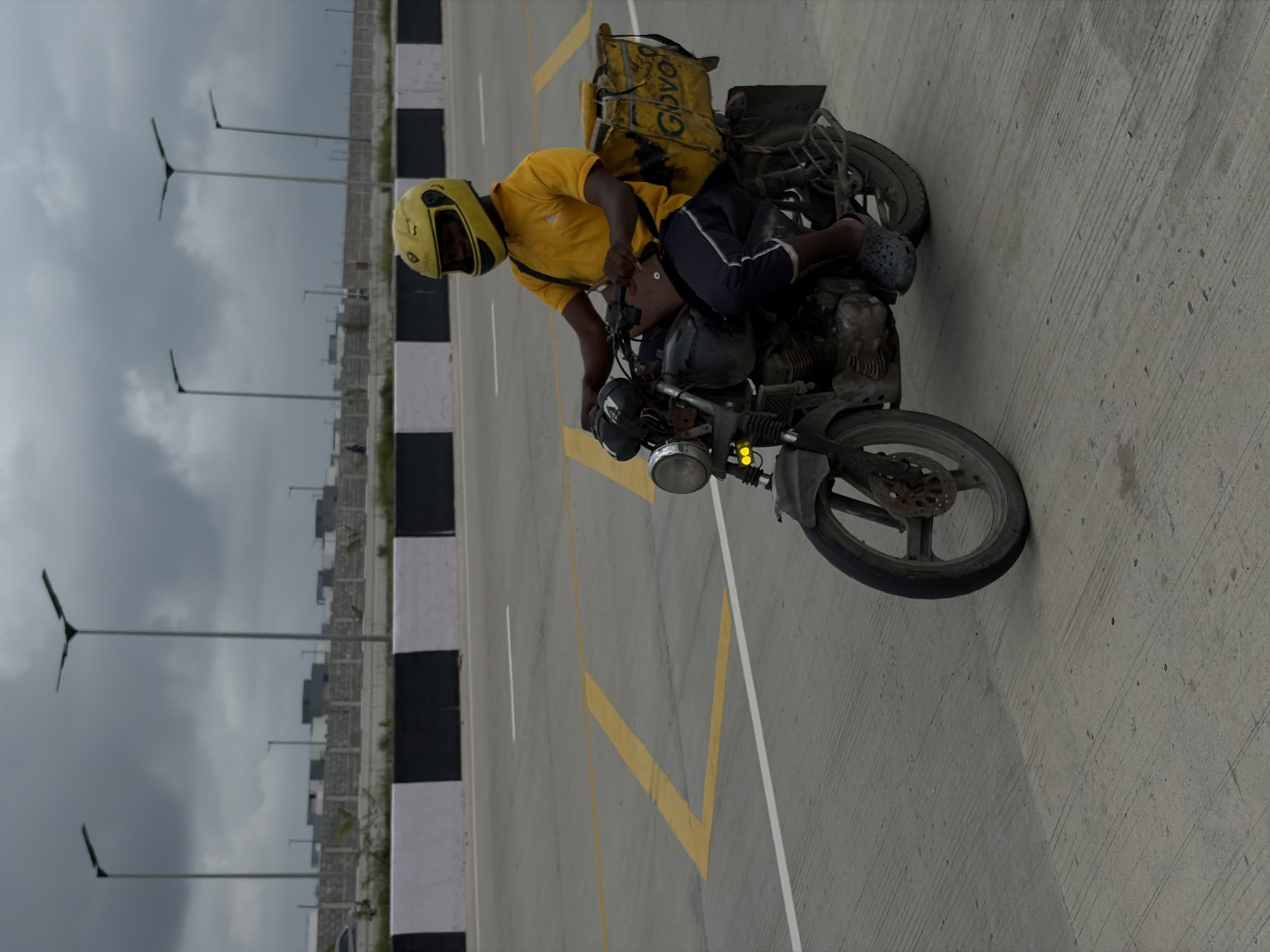 A delivery person on a motorcycle rides along a paved road in Igboefon, Nigeria. The rider, wearing a yellow helmet and yellow polo shirt, has a large yellow delivery bag attached to the back of the motorcycle. The background shows a construction site with several buildings under development and streetlights. The road has yellow and white markings. The sky is overcast.
