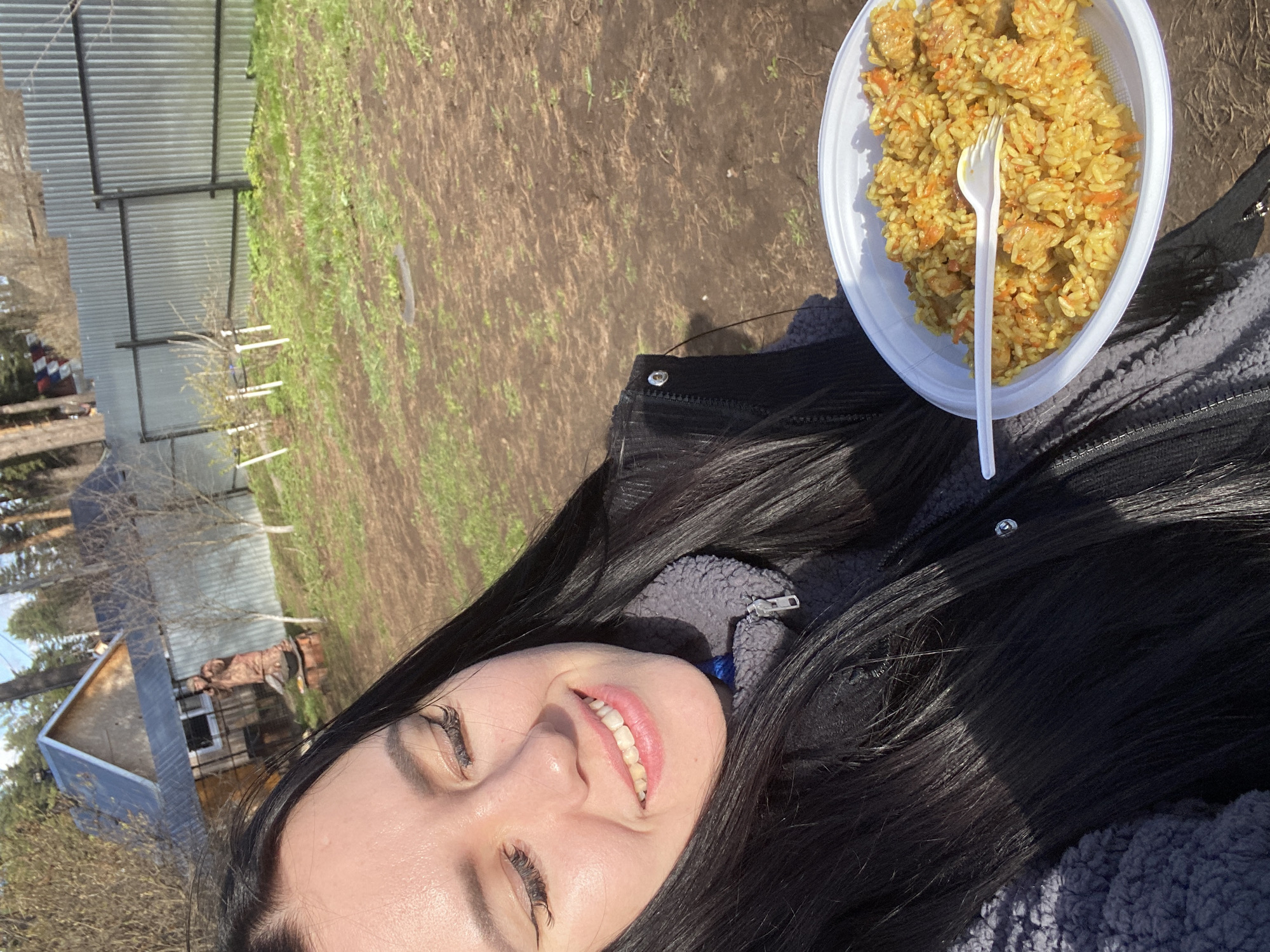 A young woman is lying outdoors, smiling at the camera. She has long, black hair and is wearing a dark jacket over a gray fleece. A plate of pilaf with a plastic fork is positioned next to her on the ground. The background includes a mix of dirt and sparse grass, with a metal fence and trees in the distance. There is a small building with a blue roof visible in the left background. Based on visual cues, the location is potentially in a rural or suburban area near the city of Chaikovsky, Russia.