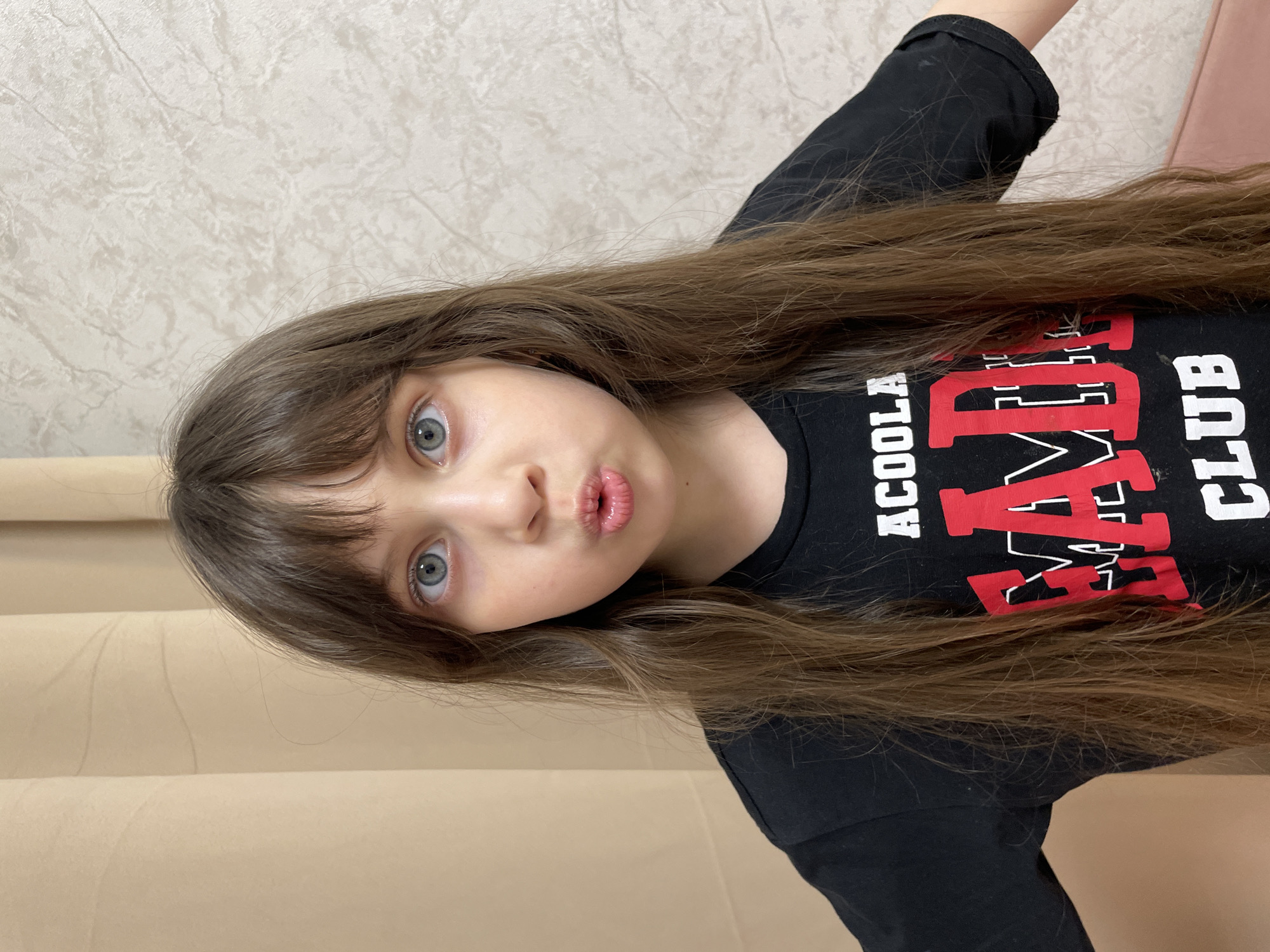 A young girl with long brown hair and bangs is posing for a photo. She is wearing a black t-shirt with a graphic design. She is looking directly at the camera with a slightly puckered expression. The background consists of a light-colored wall and a partially visible beige surface. The location is unknown.