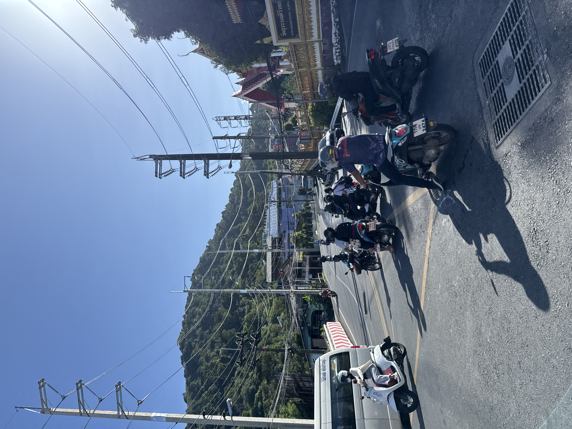 In Patong, Thailand, a traffic scene unfolds on a sun-drenched street. Several individuals are riding motorcycles and scooters, some in a queue. The road is paved, with visible yellow and white lane markings and a storm drain cover. A white van is parked on the left side of the road. In the background, a lush green mountainside rises, dotted with buildings and an abundance of power lines and utility poles. A temple with distinctive red and gold architectural features is visible on the right side of the image, with Thai text on a sign that reads "SUMANKEEREWONG TEMPLE". The sky is clear and blue.