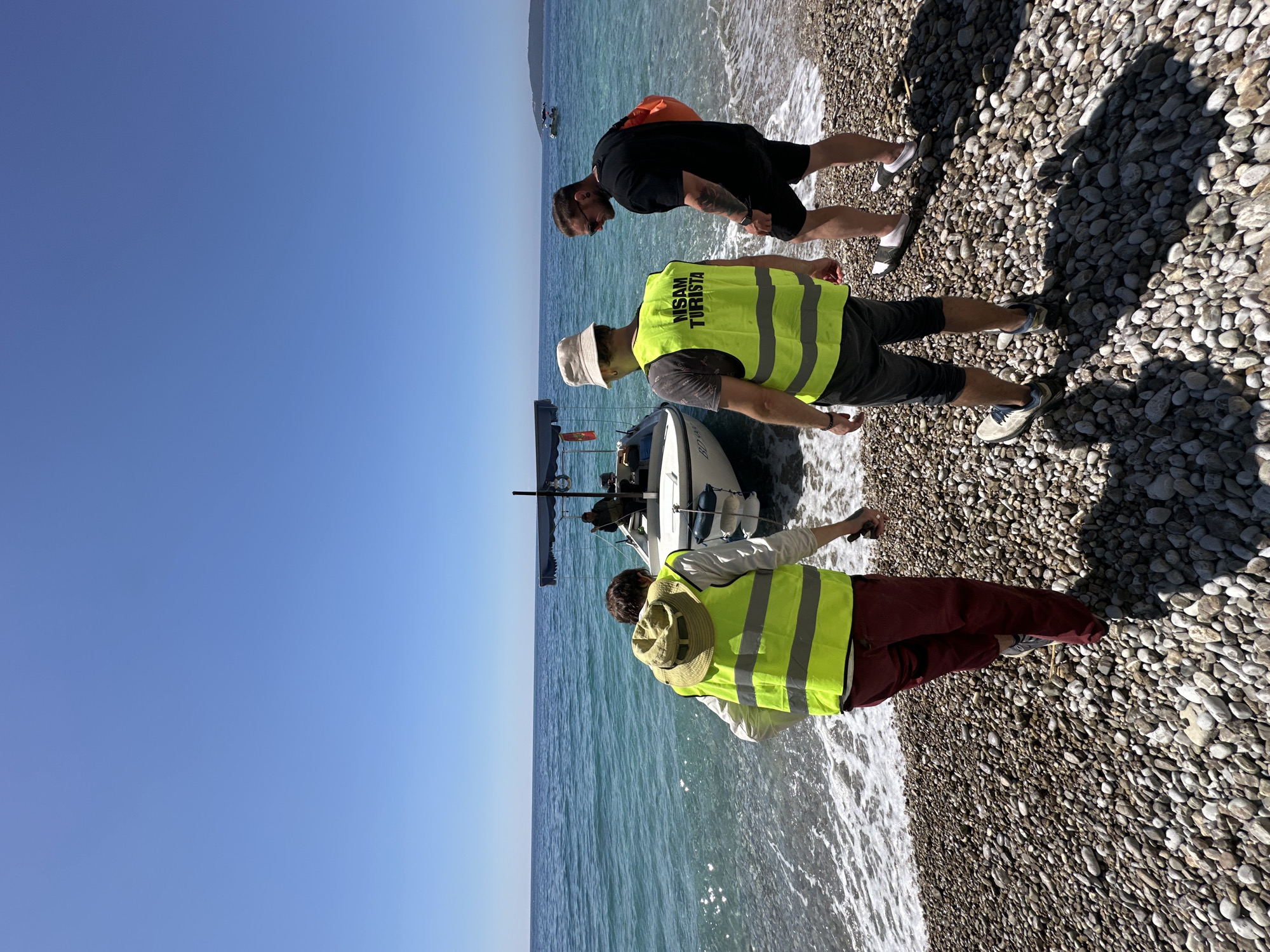 Three people are on a pebble beach beside a small boat in Budva, Montenegro. Two people are wearing lime green safety vests labeled "MSAM TURISTA". One person is wearing black shorts and a black shirt. The water is turquoise. The sky is clear and blue.