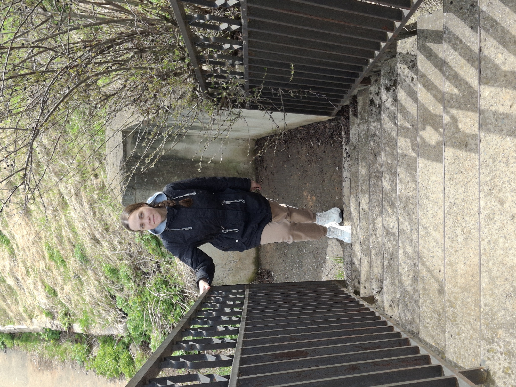An outdoor scene features a young individual standing on a concrete landing of a descending staircase. The person, with long braided brown hair, wears a black hooded jacket over a light blue hoodie, light brown trousers, and white sneakers. They gaze directly at the camera with a neutral expression, with one hand resting on the dark metal railing to their right.
The staircase is made of concrete steps and features dark metal railings with horizontal bars on both sides. To the left of the stairs, a steep bank is covered in dry grasses, shrubs, and bare tree branches, some showing early signs of green foliage. The area where the person stands is a mix of concrete and dirt. Bright sunlight illuminates the scene, casting prominent striped shadows of the railing onto the concrete steps, indicating a clear, sunny day, likely late morning or afternoon. The sparse vegetation suggests either early spring or late autumn. There is no visible text. The setting appears to be a natural or semi-natural area with pedestrian access, located in Sarapul, Russia.