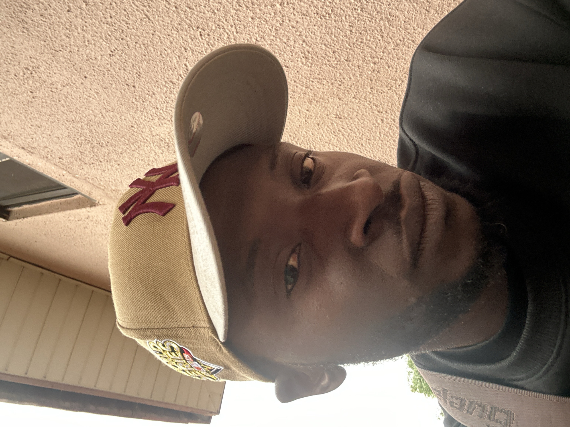 A close-up, downward-angled photograph depicts a dark-skinned man wearing a tan New York Yankees baseball cap and a black sweatshirt. The man is looking upwards toward the camera. The background features a textured beige ceiling. The location is potentially inferred based on the request, but this is not explicitly stated in the image. No specific actions or interactions are discernible.