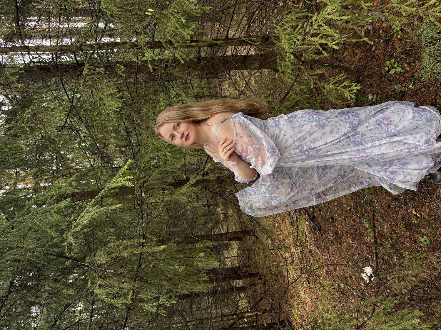 A young woman with long blonde hair and fair skin is pictured posing in a dense coniferous forest in Chaikovsky, Russia. She is wearing red lipstick and looking directly at the viewer with a neutral expression. She is dressed in a flowing, light-colored maxi dress adorned with prominent purple and blue floral patterns, featuring sheer, puffy sleeves and delicate straps. The woman is positioned in a semi-kneeling or seated posture on the forest floor, with her body angled and her arms crossed gently in front of her chest.
The setting is characterized by numerous tall, slender coniferous trees with dark trunks and vibrant green needles, suggesting a mature and lush forest. The ground is covered with a carpet of brown pine needles, fallen leaves, and some sparse green foliage. The lighting is natural daylight, appearing soft and diffused, possibly due to an overcast sky or the dense tree canopy, which creates an even illumination without harsh shadows. The overall ambiance is tranquil and natural. No visible text is present in the image. The image is oriented sideways, with the woman appearing to lie or lean, but her pose suggests she is seated or kneeling on the ground.