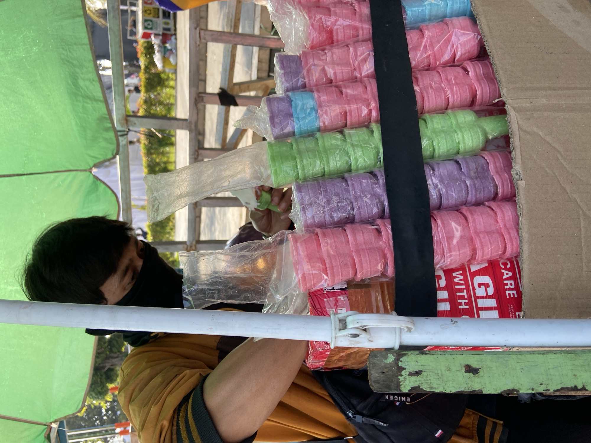 In Kecamatan Pati, Indonesia, a street vendor wearing a black face mask and a yellow jacket is arranging colorful ice cream cones. The vendor is under a large green umbrella, providing shade. The ice cream cones are stacked in rows and come in various pastel colors including pink, purple, green, and blue. They appear to be packaged in clear plastic. Behind the vendor, a wooden cart with more colorful items and possibly other goods is visible. The scene suggests a bustling outdoor market or street-side stall.