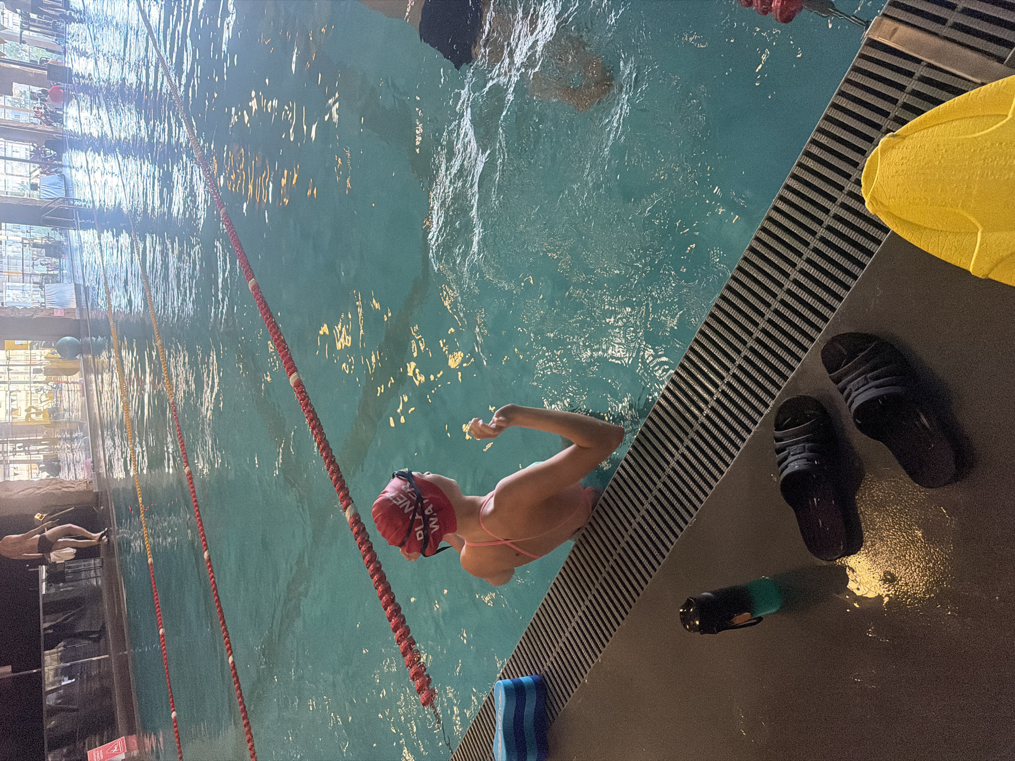 The image shows a swimming pool with lane ropes. In the foreground, there are black flip-flops, a black and green water bottle, and a yellow swim fin on the pool deck. A person wearing a red swim cap and a pink swimsuit is in the water, near the edge of the pool, facing away from the camera. Another person, mostly obscured by the water and shadows, is visible in the background, standing at the edge of the pool. The pool area appears to be indoors, with lighting reflecting off the water's surface, and a rocky texture on the far wall. The overall impression is of a public swimming facility, likely in an urban environment. The location is the city of Kyiv, Ukraine.