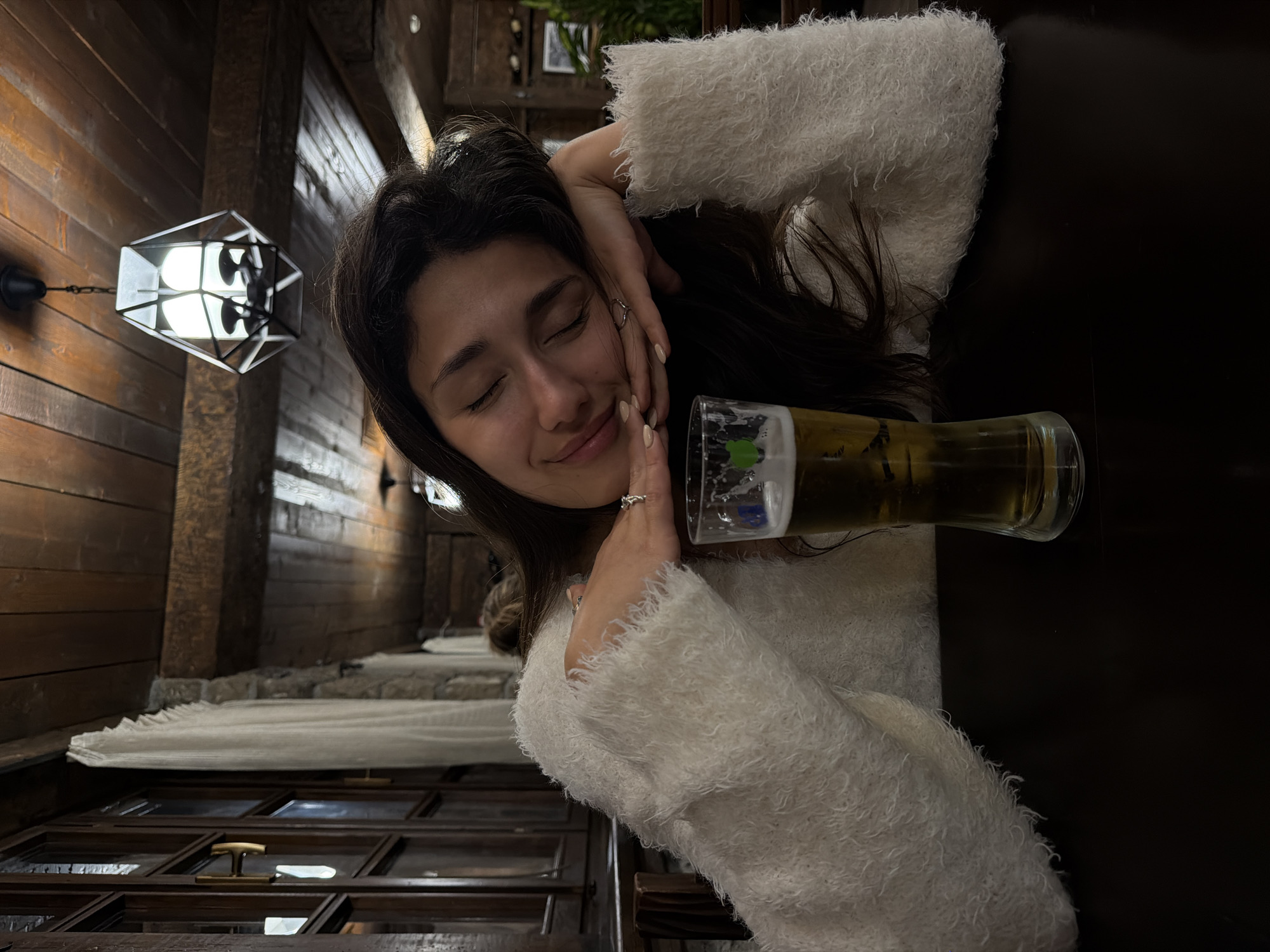 The image depicts a young woman in an indoor setting, appearing relaxed and content. She has her eyes closed and a soft smile, with her dark hair framing her face. She is wearing a light-colored, fluffy sweater or top. Her hands are gracefully positioned near her face, and a ring is visible on one of her fingers.
Beside her, a tall glass filled with an amber-colored beverage, likely beer, with foam at the top, rests on what appears to be a dark table or surface.
The background features rustic wooden paneling on the walls, suggesting a cozy or traditional interior, possibly a restaurant or pub. A distinct, geometric-shaped light fixture with exposed bulbs hangs on the wooden wall, providing artificial illumination. Further down, elements that resemble a window or a dark, framed structure with horizontal elements and possibly a curtain are visible. The overall ambiance is warm and inviting, illuminated by the artificial light.
The woman's closed eyes and gentle smile convey a feeling of peace and enjoyment, as if savoring the moment or posing for a picture in a state of calm. No visible text is clearly discernible in the image.