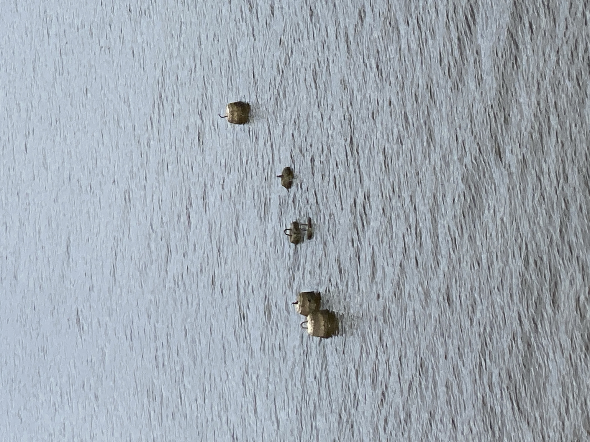 The image shows a body of water, likely a river or lake, with several cylindrical concrete objects, possibly mooring buoys, partially submerged and protruding from the surface. There are five such objects visible in a scattered formation across the frame. Three of the objects are closer to the left side of the image, while the other two are further to the right and slightly further back. Each object has a U-shaped metal handle or loop attached to its top. One of the buoys on the right has a dark, thin object arching upwards from its center, possibly a piece of debris or a thin branch. There appear to be two small birds perched on top of two separate buoys, one on the closest buoy on the left, and another on the furthest buoy on the right. The water is calm with a gentle ripple pattern covering the surface. The lighting suggests an overcast or diffused daylight condition. The overall scene is devoid of any identifiable structures or landmarks that would specifically indicate a location within the city of Leninsky, Russia, other than the presence of buoys which are common in bodies of water utilized for maritime or recreational purposes.