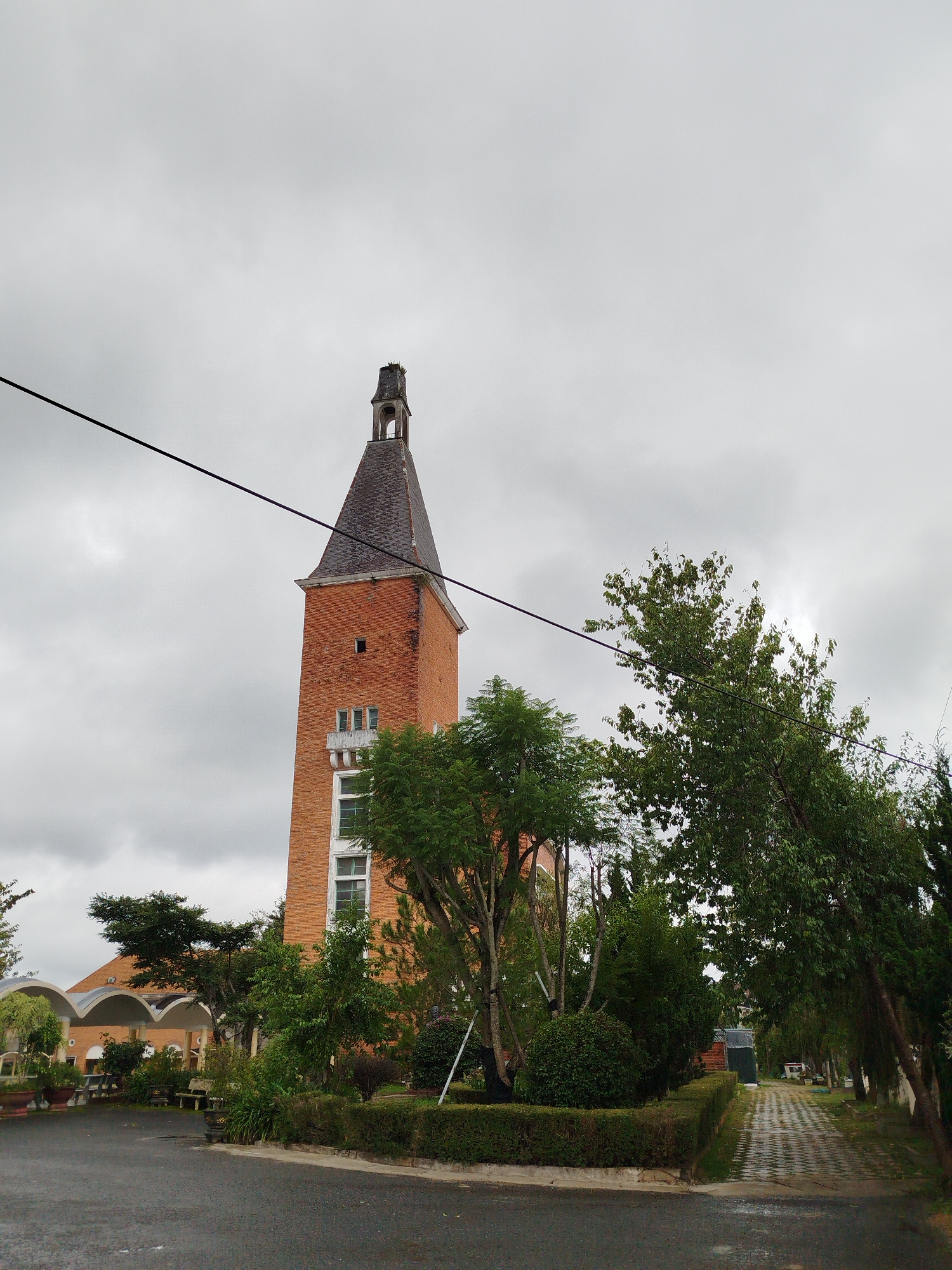 The image shows a tall, brick tower with a dark, tiled, conical roof. The tower has several windows, some of which are framed with white trim. Below the tower, there is a building with arched entrances and a tiled roof. The area in front of the buildings is a paved walkway and a dark asphalt road. There are numerous trees and manicured bushes throughout the scene. The sky is overcast and gray, indicating cloudy weather. A black power line runs diagonally across the upper part of the image. The setting appears to be a public space or a landscaped area within the city of Xuân Hương - Đà Lạt, Vietnam, possibly a historical site or tourist attraction. There are no people visible in the image.