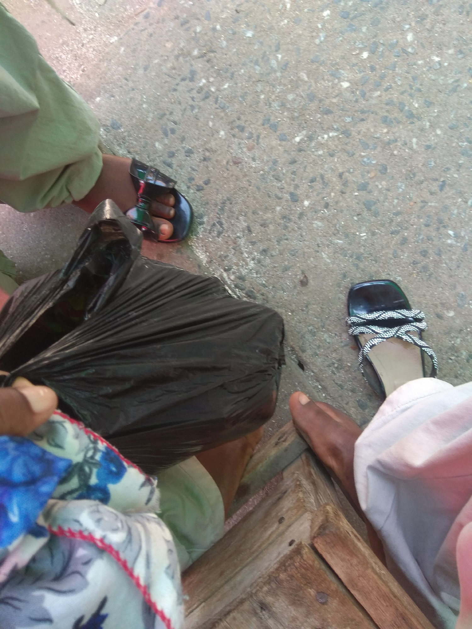 The image depicts a close-up, ground-level view of two people's feet and legs on a concrete surface, possibly in the city of Jalingo, Nigeria. To the left, one person's foot is wearing a black sandal with a patterned strap, and they are wearing light green pants. Next to this foot is a large, crumpled black plastic bag. To the right, another person's foot is wearing a black sandal with a decorative, woven white strap. This person is wearing light-colored pants, likely white or light pink. The background is a rough, speckled concrete surface with scattered small stones. In the lower right corner, the edge of a wooden structure, possibly a bench or table, is visible. The lighting suggests it is daytime.