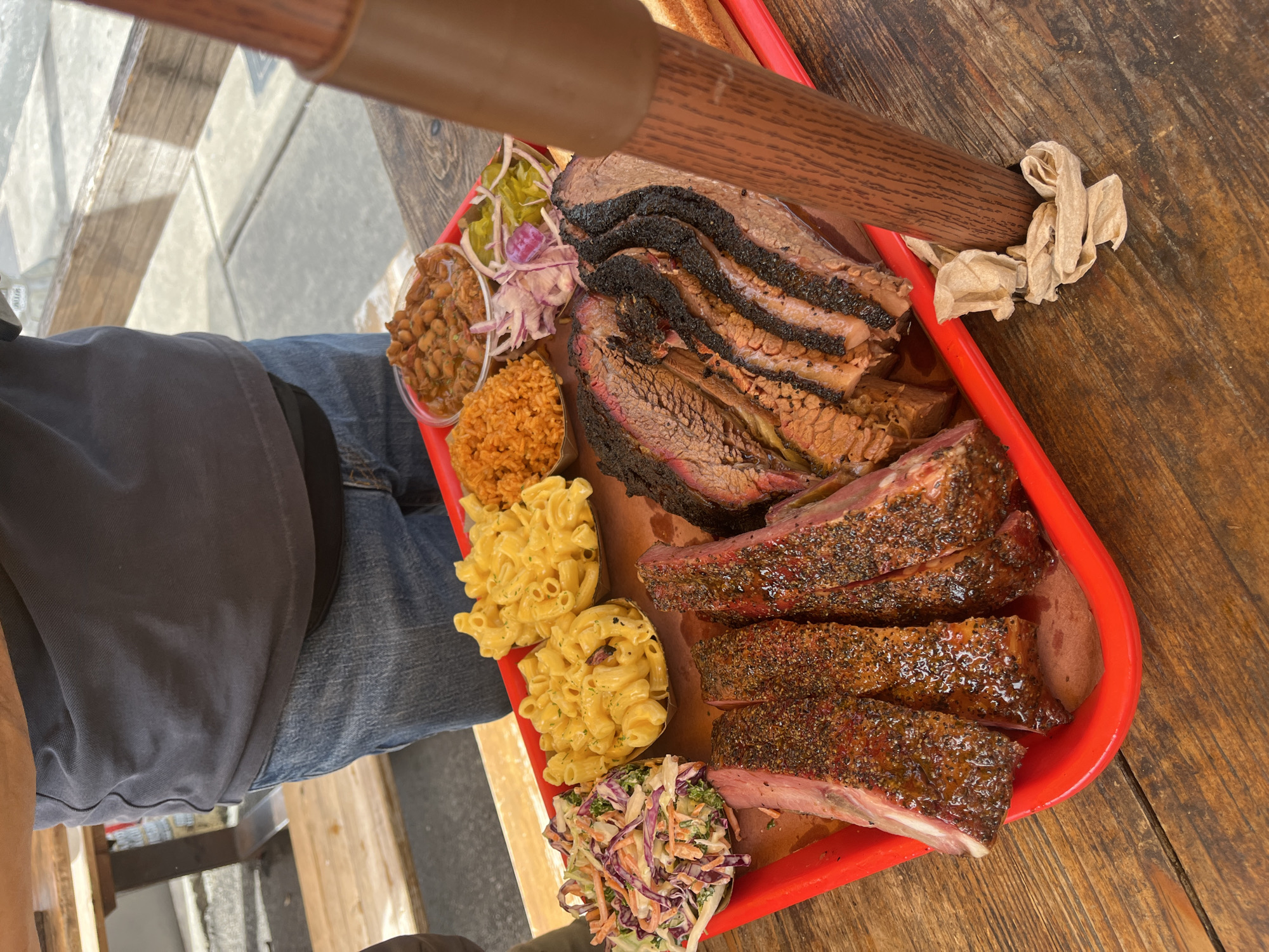 A person is holding a red tray laden with Texas barbecue. The tray is rectangular with a handle extending towards the top of the image. The food includes sliced brisket with dark bark, ribs, macaroni and cheese, rice, coleslaw, baked beans, and pickled onions and pickles. The food is arranged neatly on the tray. The person holding the tray is wearing a gray shirt and blue jeans. The tray is sitting on a wooden table. The setting appears to be an outdoor dining area, likely in a barbecue establishment. While the exact location is not discernable from the image, the type of barbecue and presentation are consistent with the style found in Austin, Texas, USA.