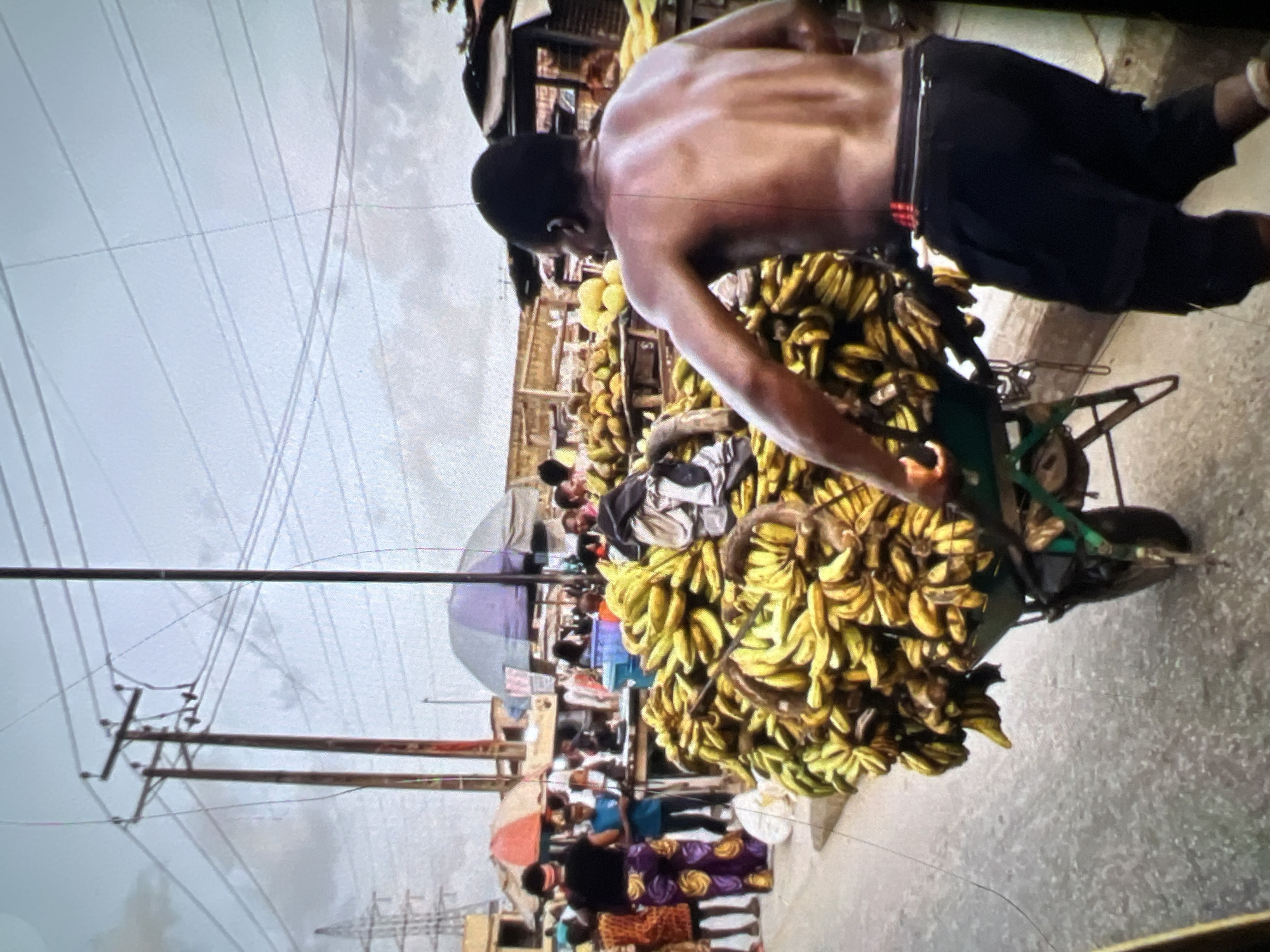In Lagos, Nigeria, a man with a bare torso pushes a wheelbarrow overflowing with bananas. The wheelbarrow is laden with a substantial amount of fruit, suggesting a large harvest or a busy market day. The man is dressed in dark shorts, and a red object is clipped to his waistband. He is walking on a paved street, likely in a bustling market area, with other people visible in the background. These individuals appear to be shoppers or vendors, engaged in various activities. The surroundings include market stalls with umbrellas providing shade, and utility poles with overhead wires, indicative of an urban environment. The sky is overcast, suggesting a cloudy day. The scene captures a moment of commerce and daily life within a Nigerian city.