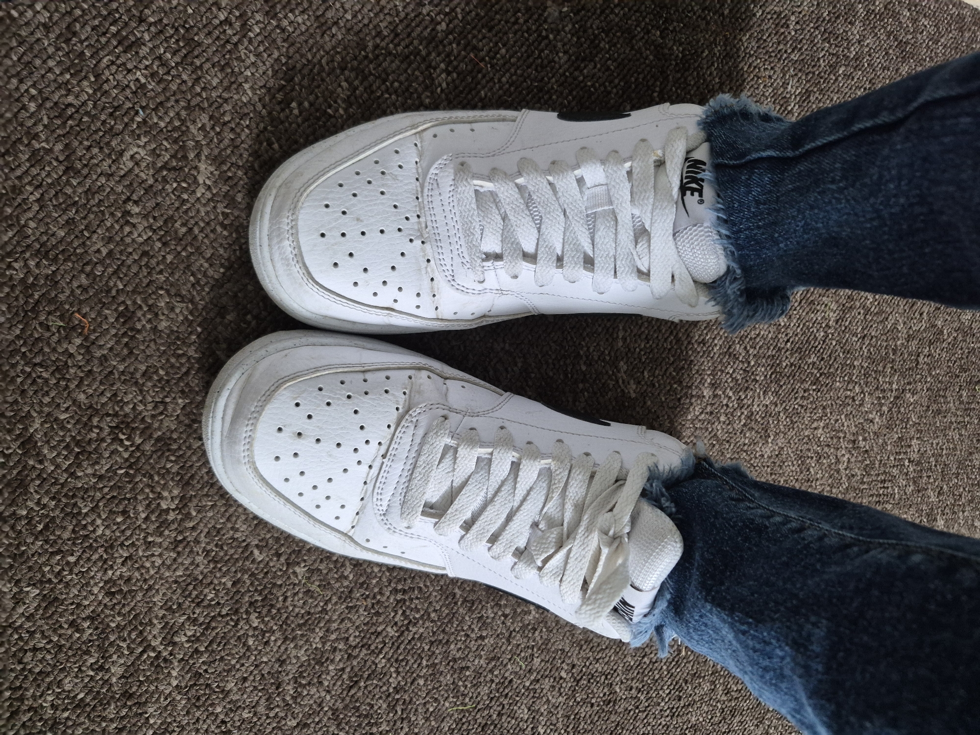 The image shows a close-up, overhead view of a person's feet wearing white Nike sneakers and distressed denim jeans. The shoes have white laces and a black Nike logo on the side. The jeans are frayed at the ankles, revealing the tops of the socks or the inside of the shoe. The shoes are standing on a brown carpeted surface. The scene appears to be indoors. There is no identifiable location in the city of Sandwell, United Kingdom.