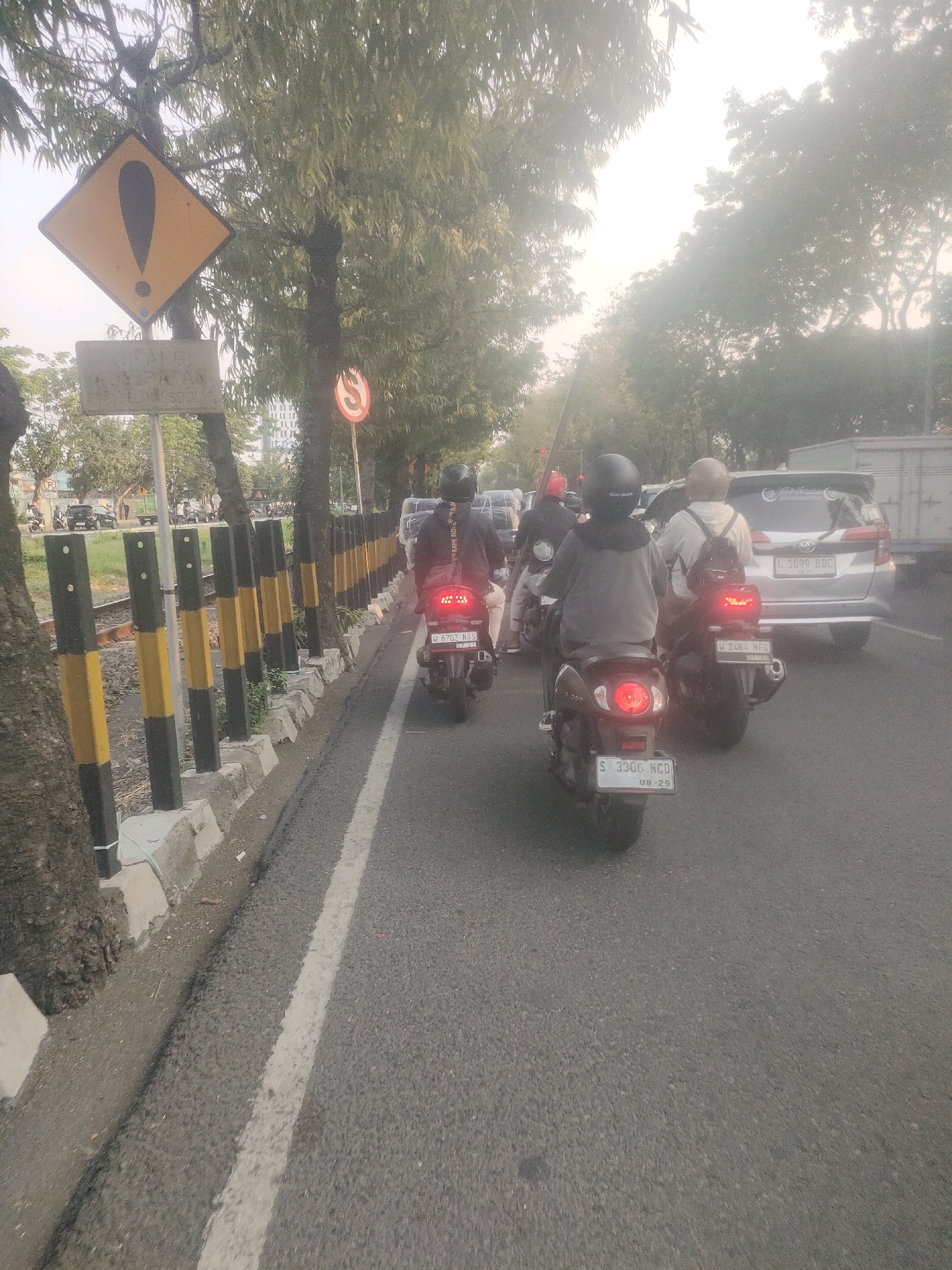 The image captures a street scene in Surabaya, Indonesia, likely during daylight hours given the lighting. The foreground is dominated by a road with a white lane marker. On the left side of the road, there's a raised concrete curb and a row of vertical bollards painted with black and yellow stripes. Beyond the bollards, a railway track is visible.
Several motorcycles are the primary subjects on the road. One motorcycle in the immediate foreground has its rear brake light illuminated and displays a license plate reading "S 3306 NCD 08-29". A rider is seated on it, wearing a helmet. Further ahead, other motorcycles are also visible, with riders in helmets. One motorcycle has a license plate "W 2464 NFC 01-29".
In the background, a silver SUV is present, with a license plate "L 1099 BDC". The environment appears to be an urban or suburban area with trees lining the roadside. A yellow diamond-shaped warning sign with a black exclamation mark is prominently displayed on a pole, accompanied by a weathered rectangular sign below it. A red circular traffic sign with a diagonal line is also visible further down the road. The overall atmosphere suggests moderate traffic and possibly slightly hazy conditions.