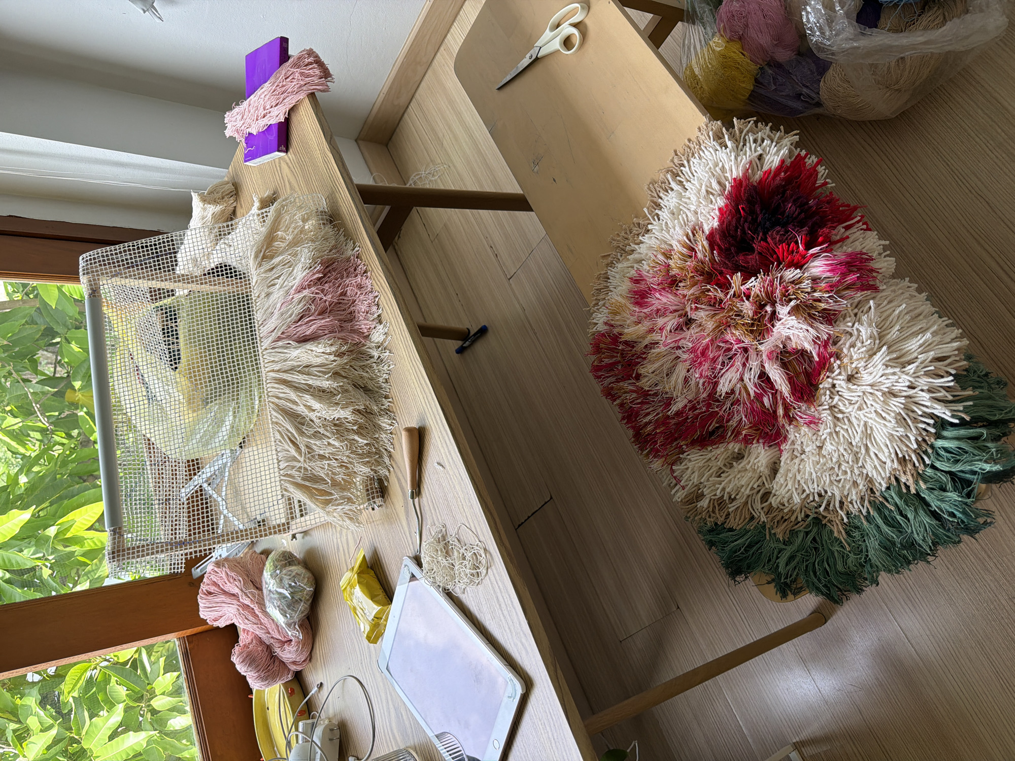 The image displays a crafting area on a wooden table, likely in Chang Phueak, Thailand, with abundant natural light from a window. A large frame, possibly for tufting or rug making, is positioned on the table, draped with a partially finished project featuring fringe in cream, pink, and beige. Various craft supplies are scattered around: balls of pink yarn, a wrapped ball of yarn, a yellow packet, a tablet, a tool with a wooden handle, and loose white yarn. To the right, a small wooden stool holds a completed rug with a vibrant, layered fringe of red, pink, and white, accented with green on the outer edges. A pair of white scissors rests on this stool. Beside the stool, a clear plastic bag contains a variety of yarn in yellow, purple, and beige colors. The floor is made of light-colored wooden planks. The backdrop features lush green foliage visible through the window, suggesting an outdoor or garden view. The overall scene conveys a dedicated crafting space in progress, highlighting a handmade rug creation.