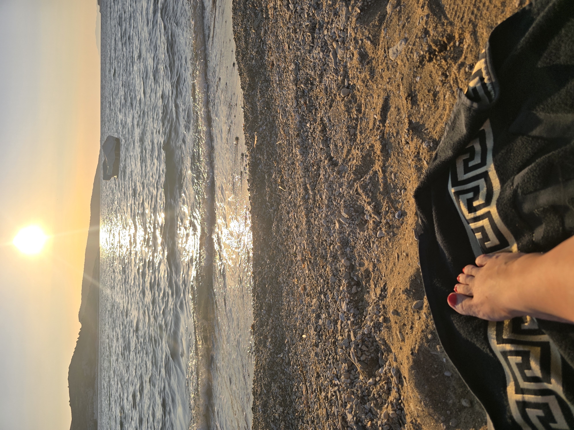 The image depicts a low-angle, ground-level view of a sandy and pebbly beach in Foça, Turkey, during a sunset. The foreground shows a person's bare foot, with red-painted toenails, resting on a black towel with a gold Greek key pattern. The towel lies on the sand, which transitions to a pebbly shore. Gentle waves lap at the edge of the beach, reflecting the warm glow of the setting sun. The sun is positioned high in the sky, casting a golden hue over the scene and creating a shimmering path on the water. In the middle ground, a small boat, covered with a grey tarp, floats on the calm sea. The background features a silhouetted hilly coastline of Foça. The overall atmosphere is tranquil and serene, capturing the essence of a peaceful evening by the sea.