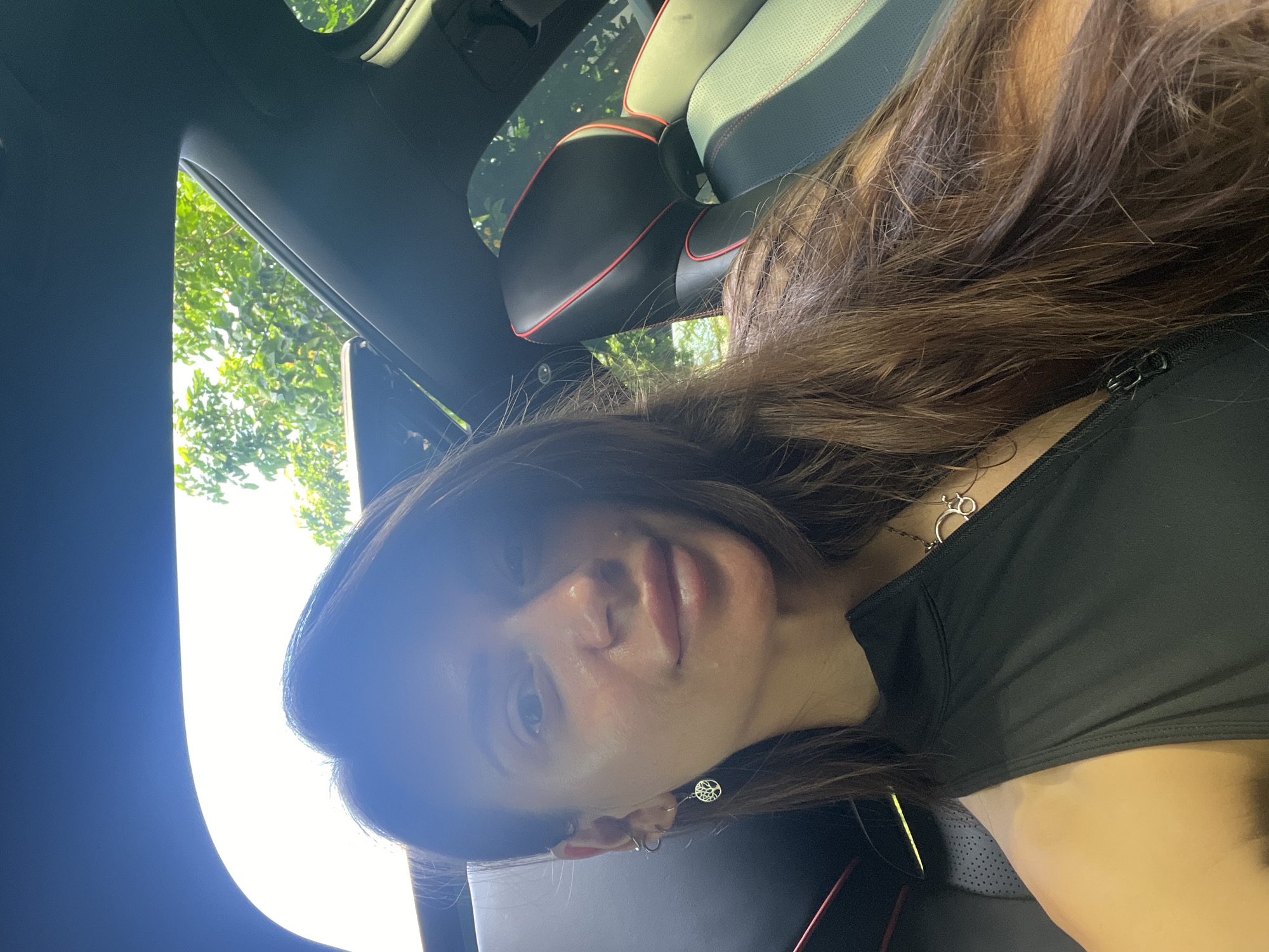 The image is a selfie taken inside a car, likely during daylight hours. The person in the photo is a woman with long brown hair, wearing a black top and a necklace. She has earrings in. The interior of the car is visible, including the dashboard, the car seat with red stitching, and a view of trees outside the window. The location or city is not identifiable from the visual information.