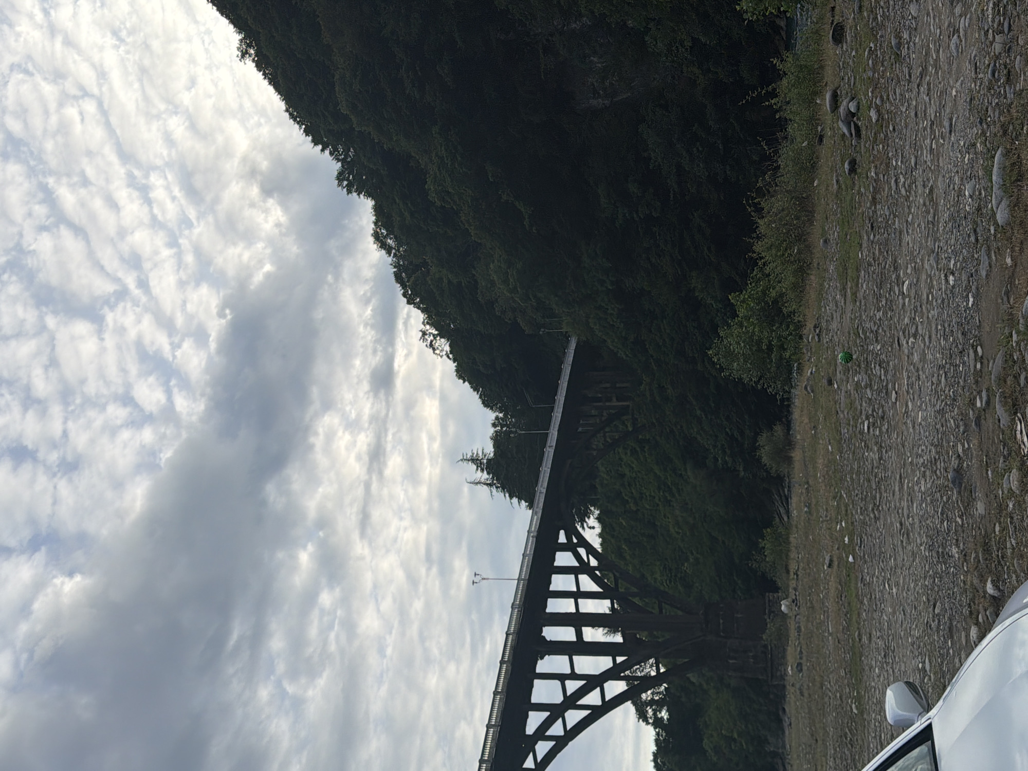 This media file captures a view of a large, arched bridge amidst a lush, tree-covered landscape. The bridge's dark, robust structure, composed of beams and supports, stands in contrast to the vibrant green foliage that blankets the steep incline behind it. The sky above is filled with a dynamic display of clouds, ranging from wispy cirrus formations to larger, more diffused grey masses, suggesting a partly cloudy or overcast day.
The scene appears to be set in a natural environment, likely a valley or gorge, given the elevated position of the bridge and the dense forestation. The ground in the foreground is a rough, gravelly surface with scattered stones and patches of dry grass, hinting at a roadside or parking area. A portion of a white vehicle is visible on the right side of the frame, with its side mirror and part of its hood in view, indicating human presence and recent activity. A small, green spherical object, possibly a ball or a piece of debris, is noticeable on the gravel.
The lighting suggests it could be daytime, possibly late afternoon or early morning, as the light is diffused and the shadows are not harsh. The overall mood is serene and natural, with the man-made structure of the bridge integrated into the wilderness. There is no visible text in the image. The scene's context of Wazabaa, Abkhazia, points to a location known for its natural beauty and potentially dramatic topography, which this image effectively portrays.