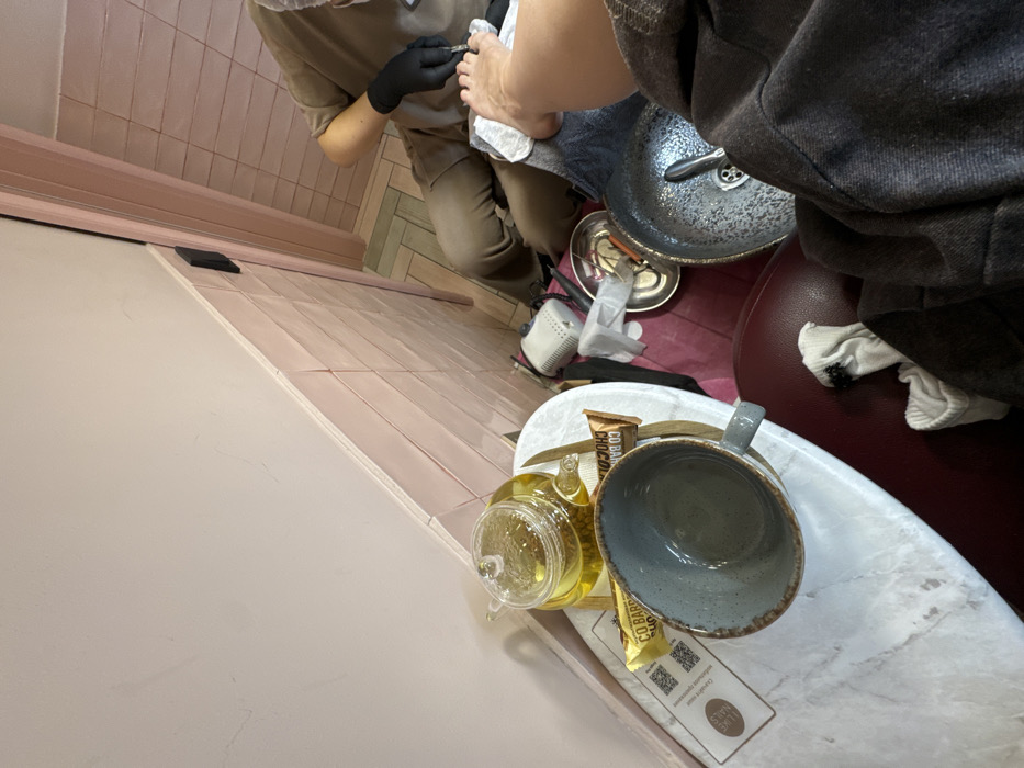 The image displays an interior scene within a nail salon or spa, specifically a pedicure station in Moscow, Russia. The perspective is from the client, looking down at their right foot and the pedicurist.
**People:**
1. A client, whose lower right leg and foot are prominently featured, resting on a white towel over a dark basin. Their left lower leg, with white socks around the ankle, is partially visible. The client wears dark, possibly grey, trousers or a robe.
2. A pedicurist, positioned in front of the client's foot, is partially visible from the torso downwards. They wear a light brown uniform, a hairnet or cap, and black examination gloves. The pedicurist is actively using a silver-colored tool to perform a service on the client's right big toenail.
**Objects:**
* A small, oval-shaped side table with a white marble-effect surface is positioned to the client's left. On it rests a clear glass teapot filled with amber liquid (likely tea), a grey ceramic mug, and two individually wrapped "CO BAR" branded chocolate bars. A white informational sign with a QR code and the text "LOVE NAILS" (in English and Cyrillic) is also on the table.
* At the pedicurist's workstation, a dark metallic basin with a drain contains water and possibly tools. Various manicure/pedicure tools, a small white electronic device, and a crumpled white tissue are arranged on a pink mat on the floor beside the basin.
**Environment and Location:**
The room's walls are characterized by light pink rectangular tiles in the lower section, with the upper portion painted a smooth, light beige or off-white color. A doorway with a pink frame is visible to the right. The floor features light-colored rectangular tiles arranged in a herringbone pattern, incorporating subtle pale green accents. The sign on the side table identifies the establishment as "LOVE NAILS" with the address "UL. SUSCHEVKY VAL, D. 5/7," indicating the specific location on Sushchevsky Val Street in Moscow, Russia.