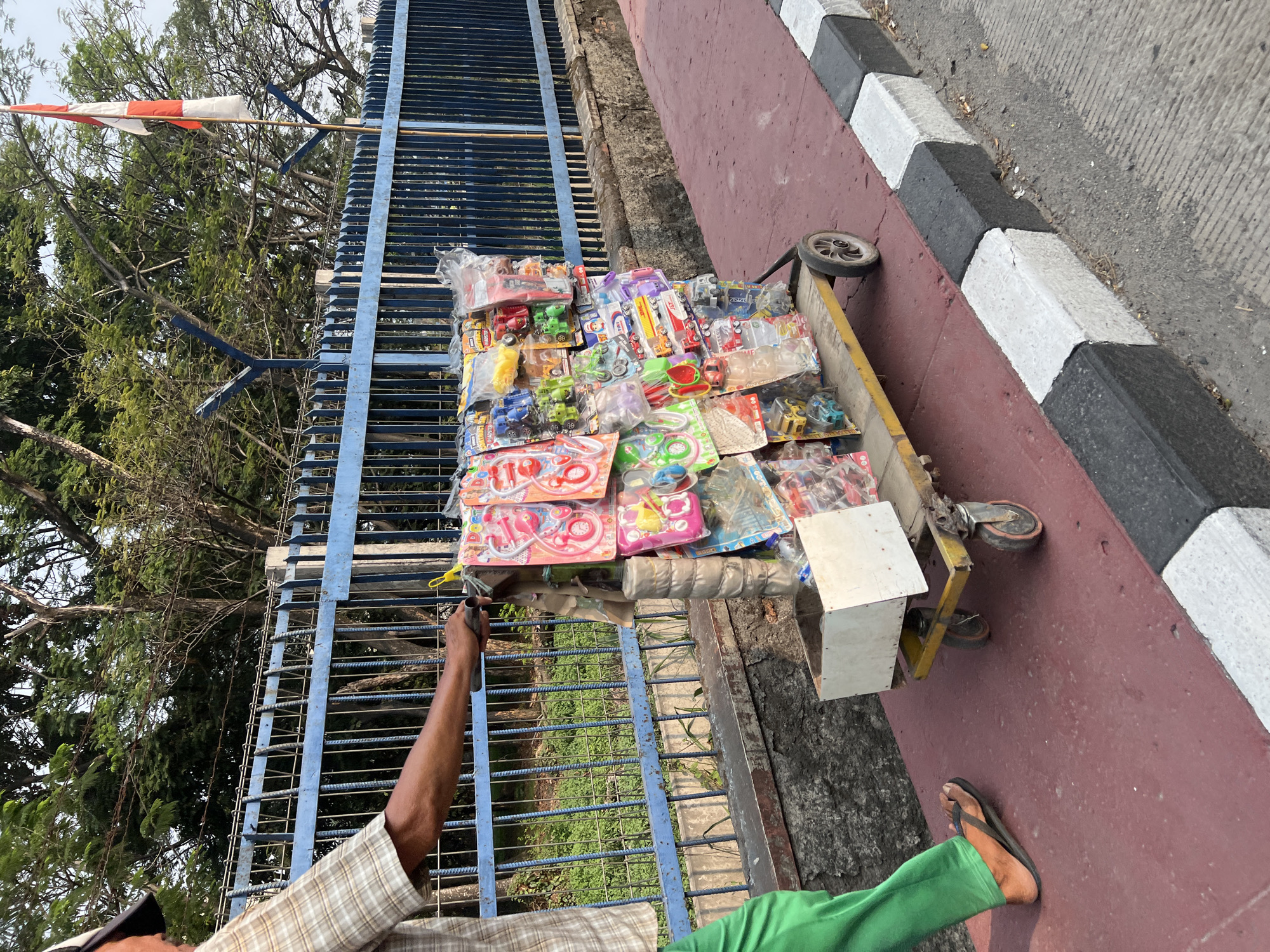 A person is pushing a cart filled with toys along a sidewalk. The cart is yellow and has four wheels. The toys are packaged and displayed in rows. The person is wearing green pants and a checkered shirt. The sidewalk is red, and there is a black and white curb along the side. In the background, there is a blue fence and trees. A flag is visible in the upper right corner of the image. This scene is likely in Citeureup, Indonesia, as indicated by the presence of a flag that appears to be the Indonesian flag and the overall environment which is consistent with urban areas in Indonesia. The person's attire and the type of cart suggest a street vendor.