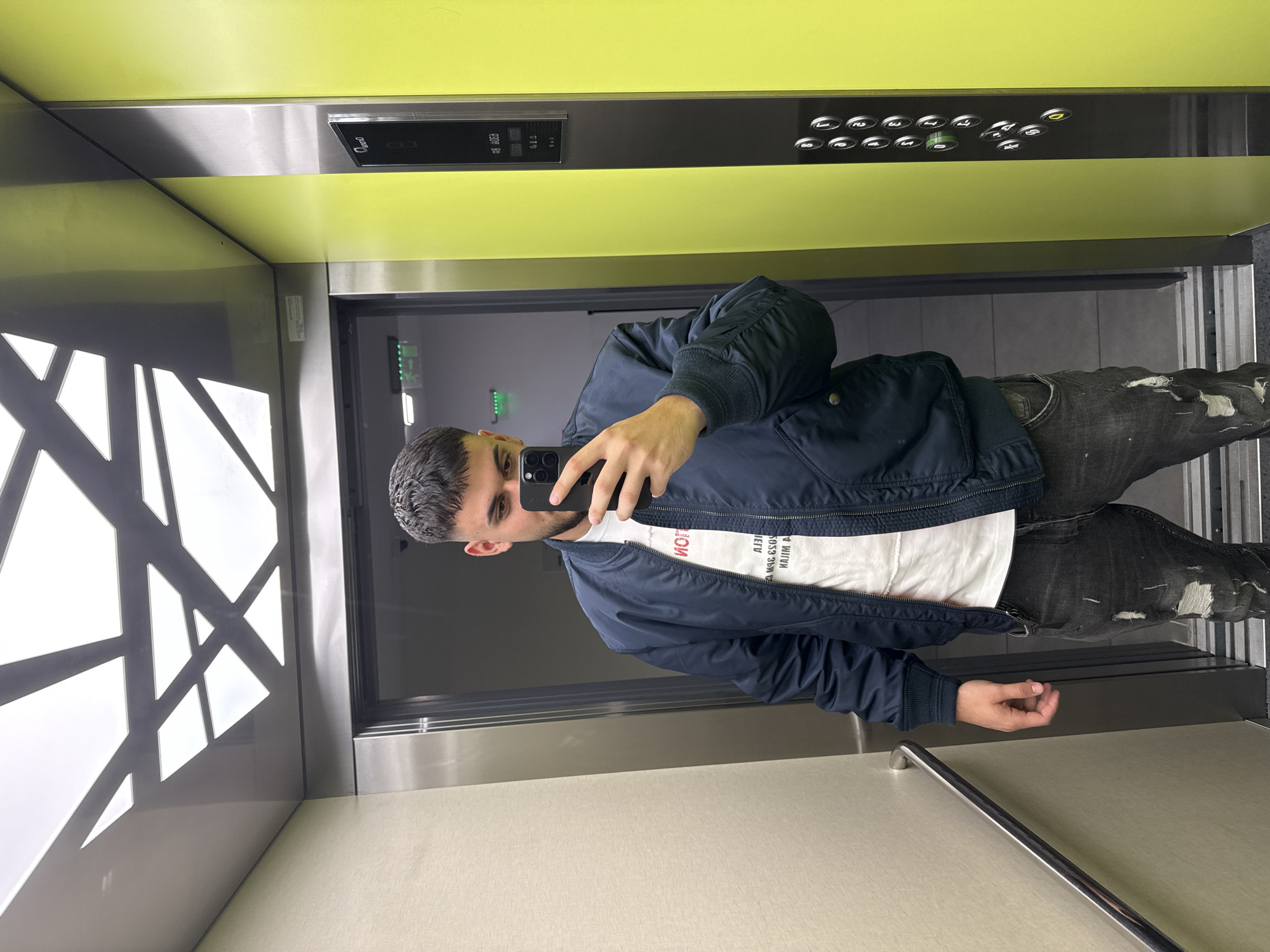A man is depicted taking a mirror selfie inside a modern elevator. He has short dark hair and a light beard, wearing a dark blue bomber jacket over a white t-shirt with mirrored text, and dark, distressed jeans. He holds a smartphone to capture his reflection, with a neutral expression.
The elevator interior features bright lime green panels on the upper walls, contrasting with metallic grey surfaces on the lower sections. On the left wall, a decorative panel showcases an abstract geometric pattern in white against a grey background. A metallic handrail extends along the right side of the elevator.
The dark grey control panel is integrated into the lime green wall, featuring numerous silver-rimmed buttons for different floors, ranging from 0 to 8, with some illuminated in green or yellow. A small rectangular screen on the panel displays "234" and "00", and the word "Door" is visible above. The mirrored text on the man's white t-shirt includes "HOTEL" and other partially obscured words and alphanumeric codes. The elevator is brightly lit, indicating an indoor setting in a modern building, possibly in Sofia, Bulgaria, as per the provided context.