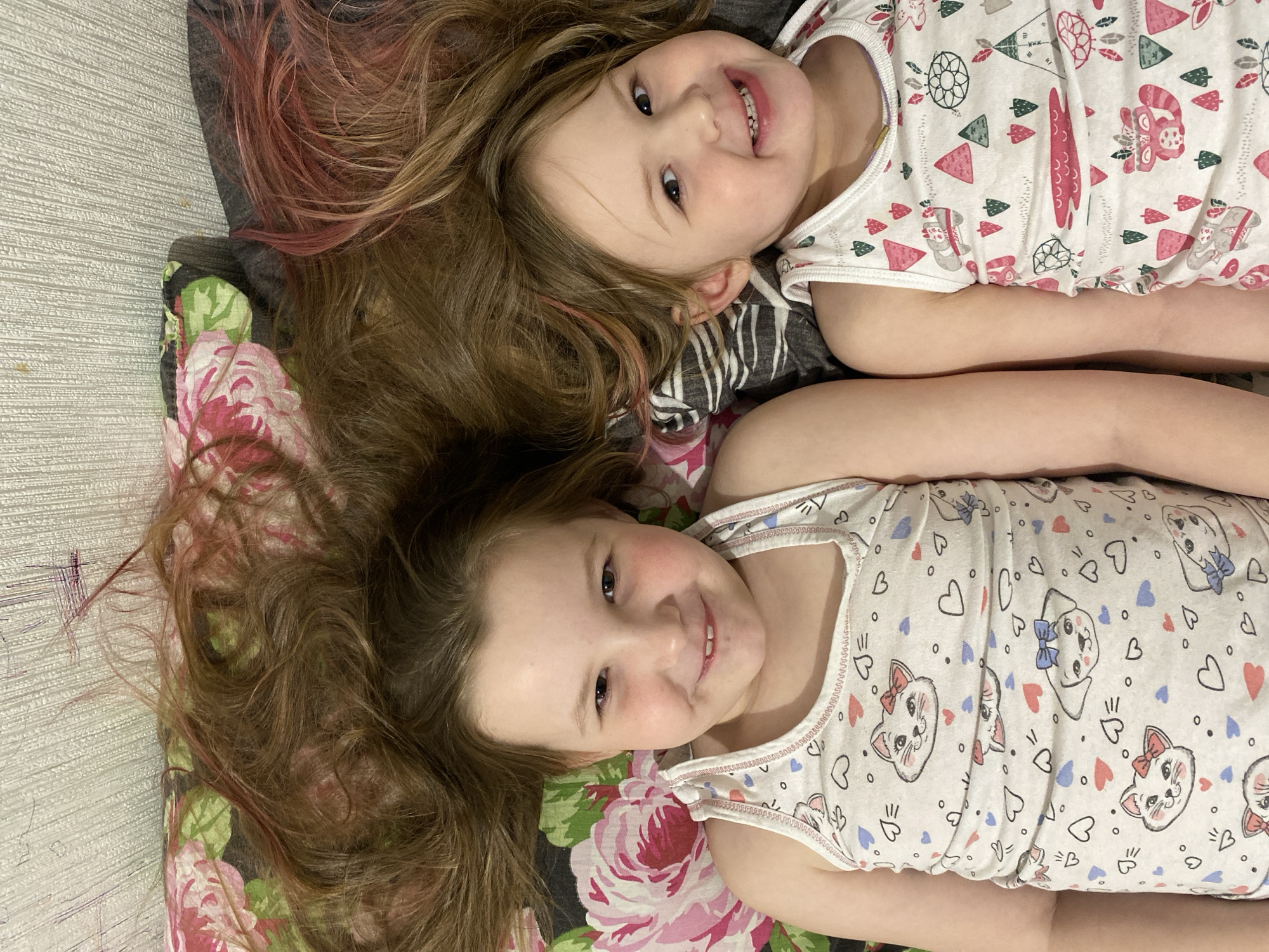 Two young girls lie side-by-side on a bed or similar surface. One girl is positioned at the top of the frame and the other at the bottom. They are both wearing white sleeveless shirts with patterns; the top girl's shirt has a mix of teepees, animals, and feathers, while the bottom girl's shirt is adorned with cat faces and hearts. Both girls have brown hair, with the top girl having pink highlights. Their heads rest on a pillow or cushion with a floral pattern of pink and green. The background includes a textured, light-colored wall or surface. The location is Chaikovsky, Russia.