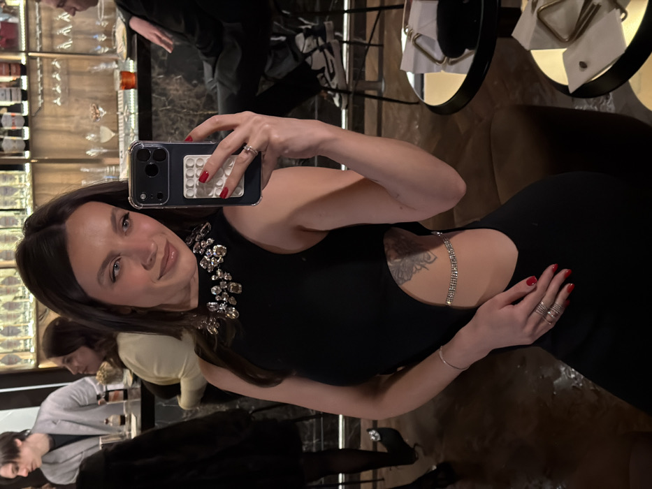 A woman in a black dress with a jeweled collar takes a selfie in a bar. The dress has a cutout at the waistline, adorned with a jeweled chain. She holds her phone, which has a clear case and a white "pop-it" fidget toy attached. Her nails are painted red, and she wears multiple rings. The background includes bar shelves with bottles and other patrons.