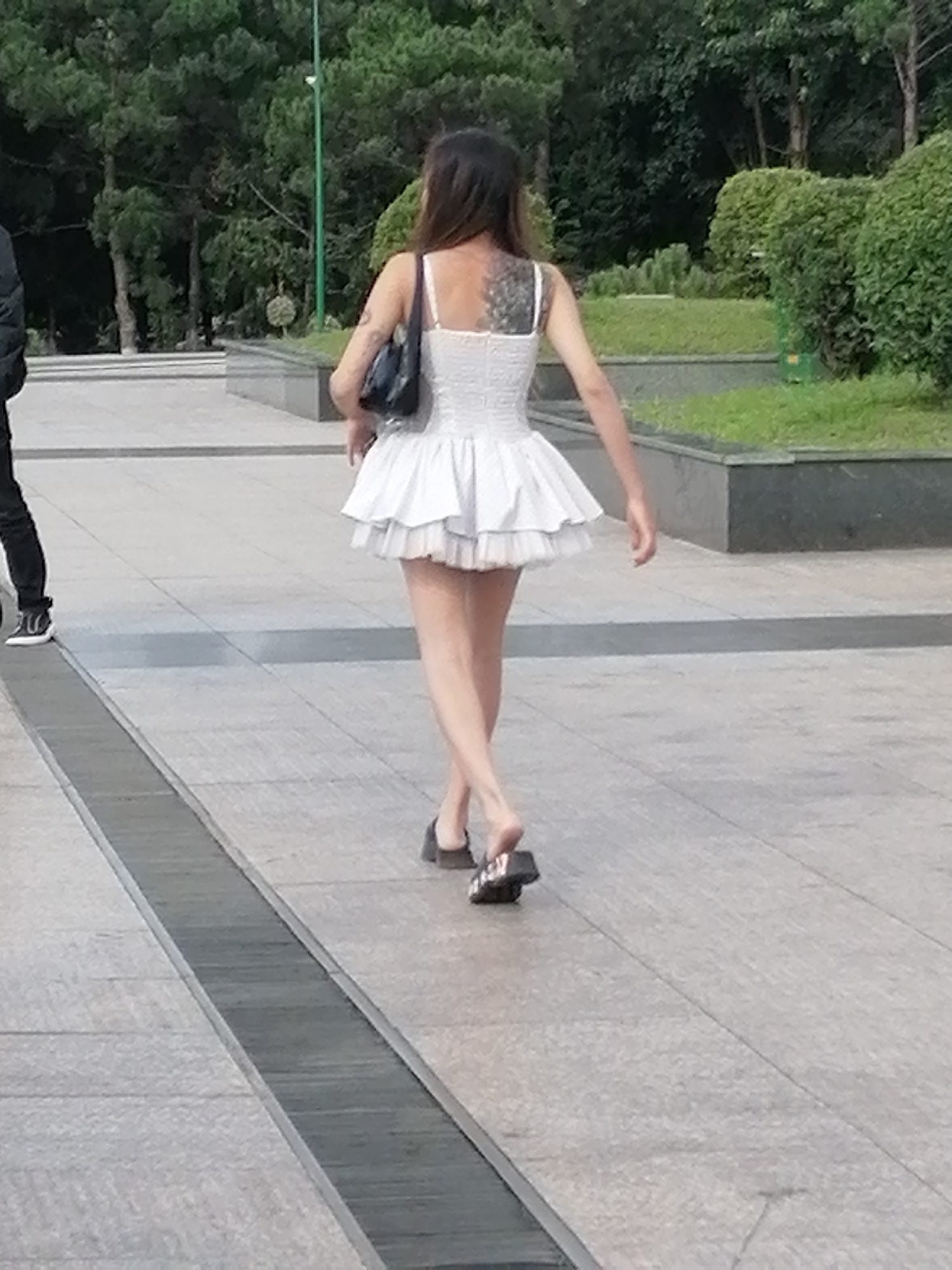A woman with dark hair and a large tattoo on her back walks away from the camera. She is wearing a white, ruffled dress and dark sandals. She carries a dark purse over her left shoulder. The woman is walking on a paved pathway. To her left, a person in black pants and white sneakers walks in the opposite direction. The background consists of trees and manicured bushes. This scene is likely at the Da Lat Flower Park in Da Lat, Vietnam, a popular tourist attraction known for its gardens and walking paths.