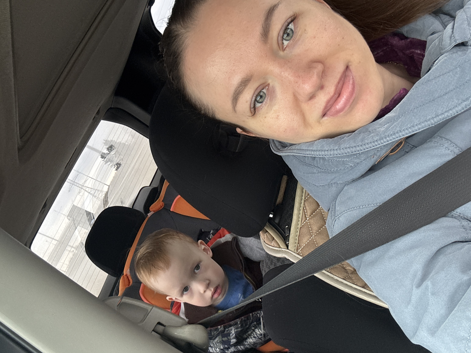 A selfie taken inside a car. A woman with light skin, green eyes, and brown hair is wearing a light blue jacket and a seatbelt. Behind her, a young boy with light hair is sitting in a car seat, also secured with a seatbelt. The background shows a building with multiple windows. The location is Lipaki, Russia.