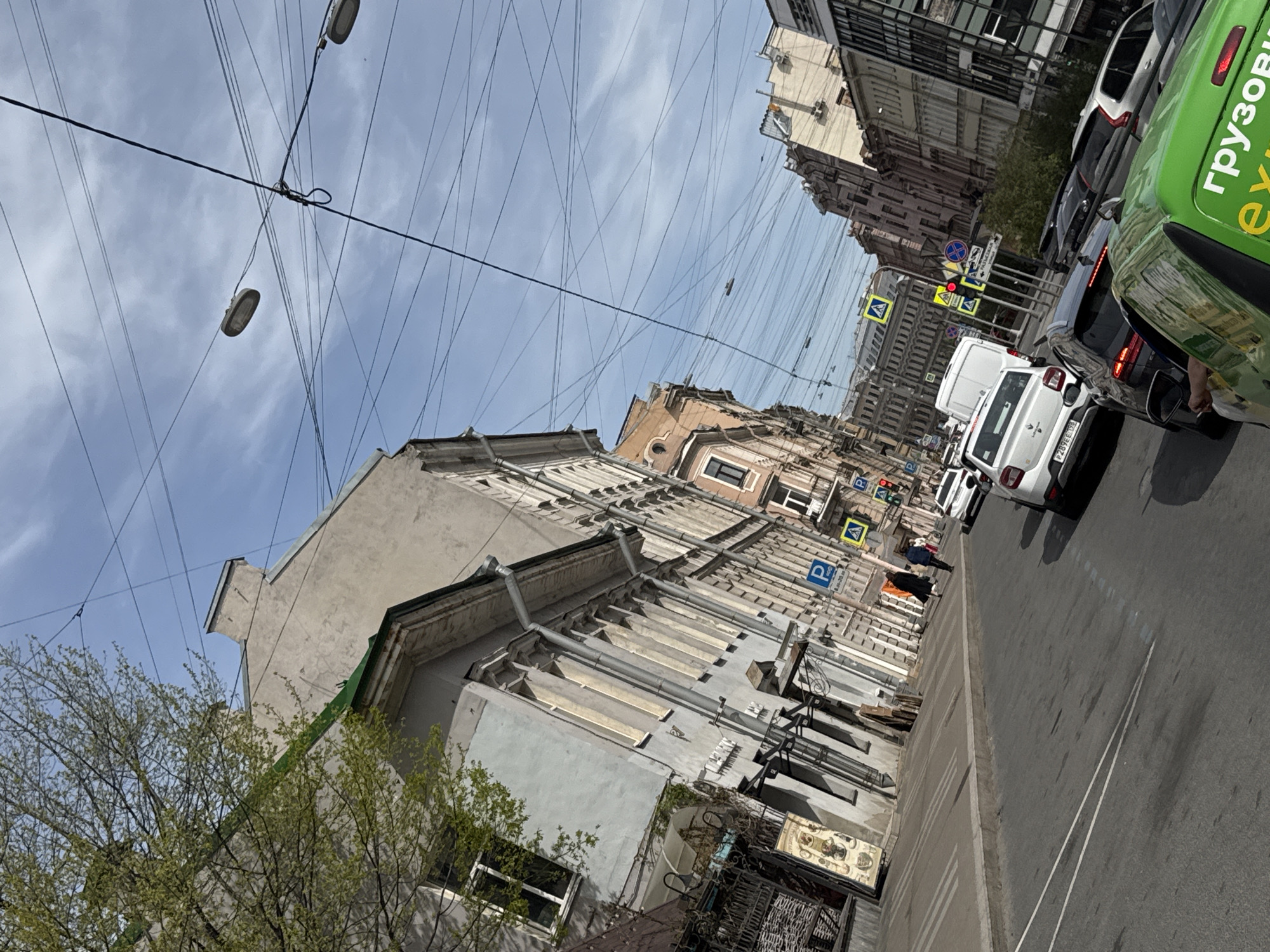 The image depicts a street scene in Saint Petersburg, Russia. Overhead, a dense network of electrical or communication wires spans the sky above the buildings. The buildings themselves are old, with gray and beige facades. Parking signs are visible on the buildings.
Traffic is present on the right side of the frame, consisting of multiple vehicles. The weather appears to be clear. Trees with budding leaves are visible in the lower left corner, implying it is springtime.
