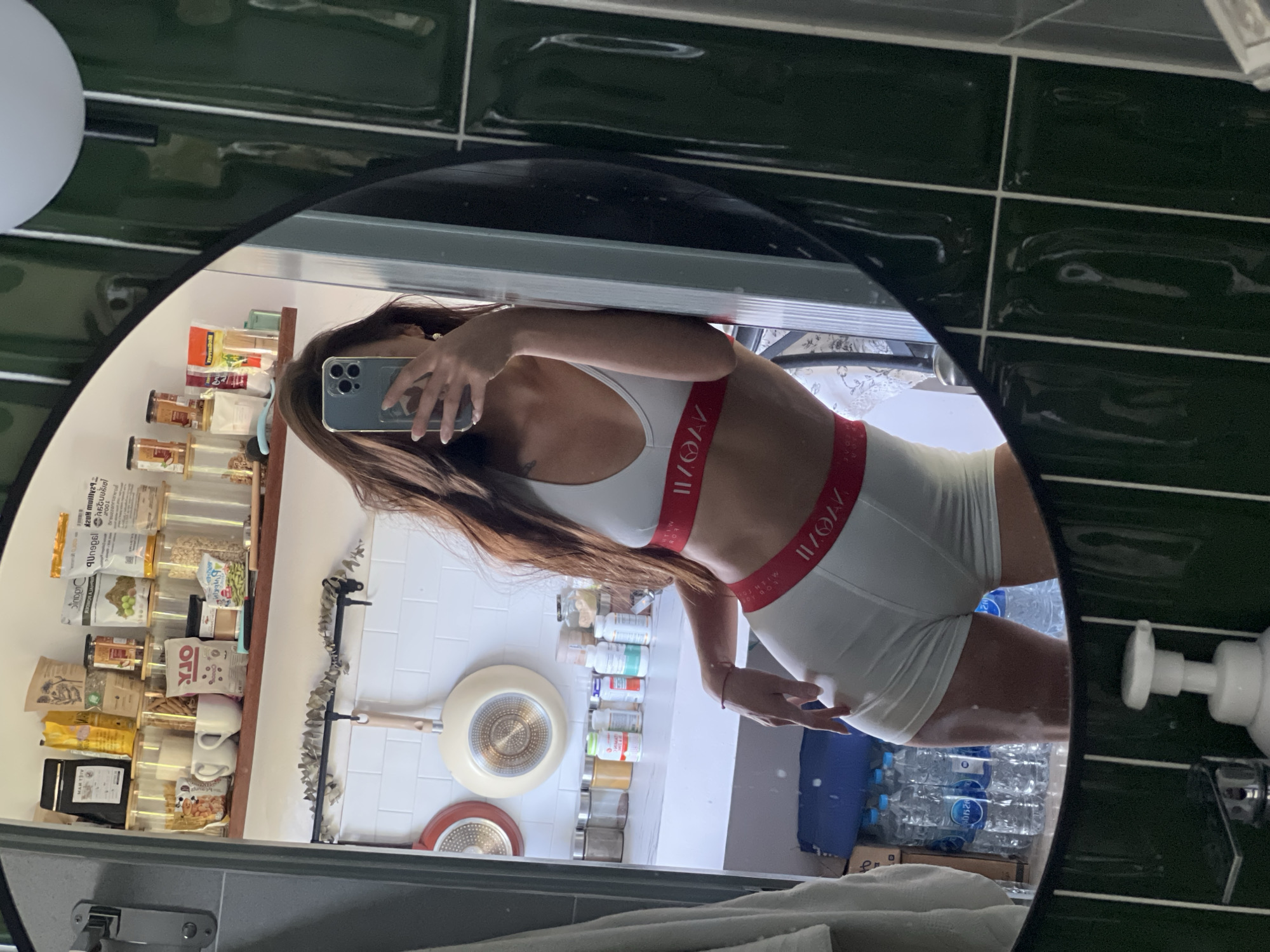 A medium, eye-level shot depicts a person facing forward in a mirror, holding a phone in their left hand. The mirror's reflection reveals shelves stocked with numerous labeled food items. A portion of the person's body, from the waist up, is visible, as well as the lower half of their legs. The clothing they are wearing includes a white top with red accents. Additional details visible in the mirror include bottles, pans, and a towel. The foreground of the shot shows the edge of a counter, while the upper right corner features green-tiled walls and a soap dispenser.