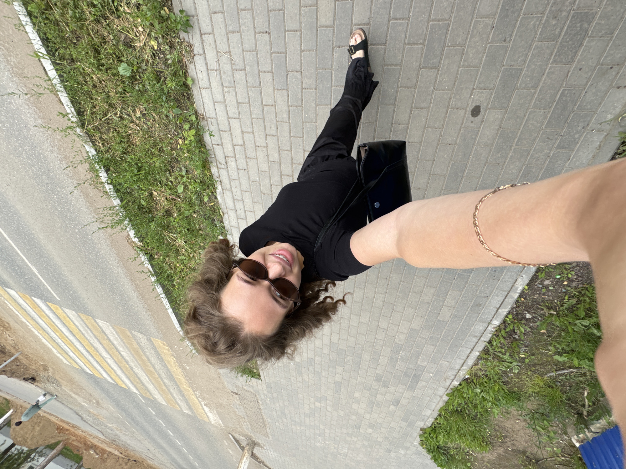 A woman with curly brown hair and sunglasses, wearing a black top and black pants, takes a selfie while standing on a paved sidewalk. She is holding a black purse. In the background, there is a road with a pedestrian crossing. The scene appears to be outdoors in an urban or suburban setting, likely in Chaikovsky, Russia, given the context.