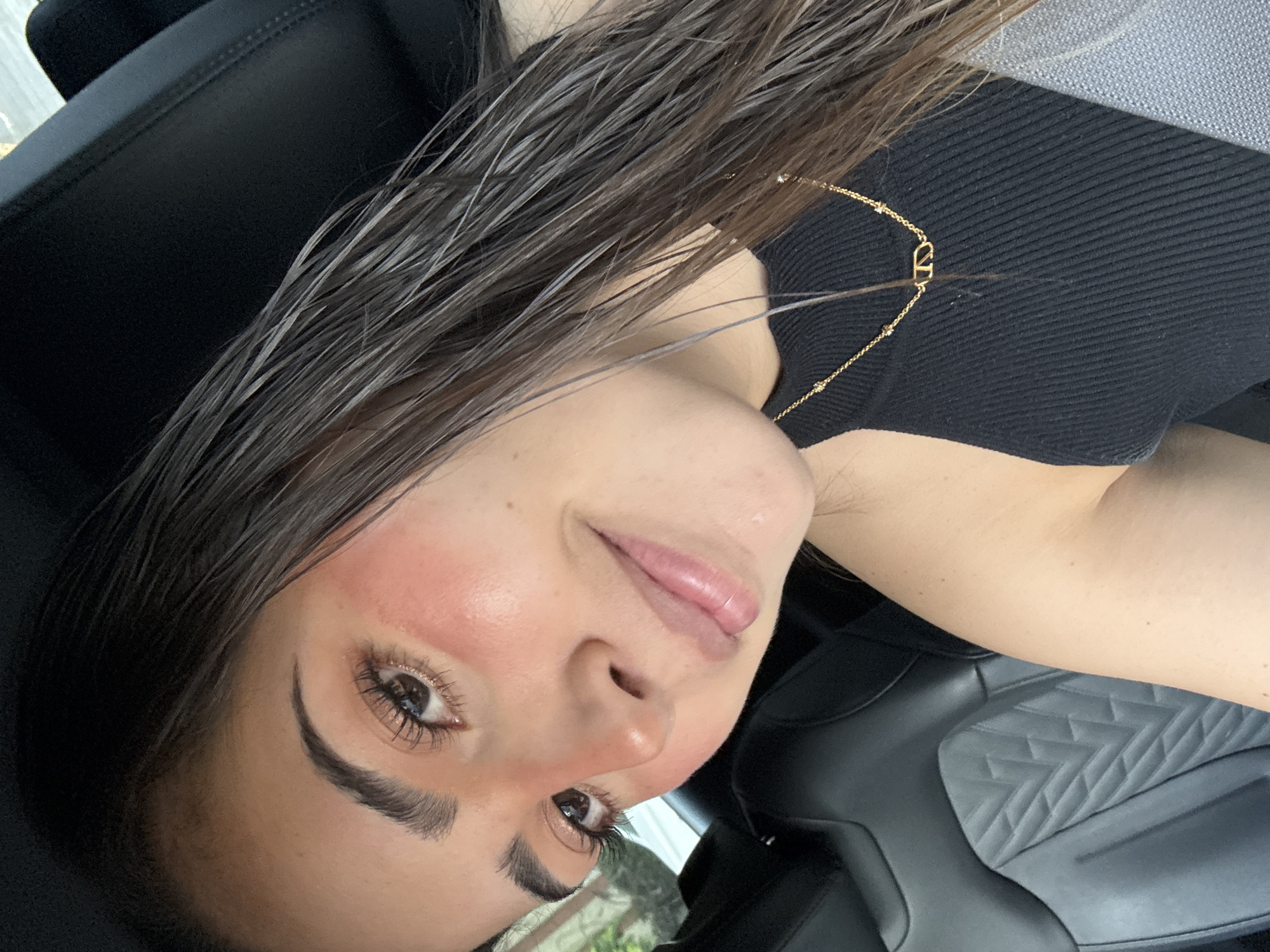 A close-up, eye-level selfie shows a young adult female positioned in the passenger seat of a vehicle. Her dark brown hair, appearing wet or styled as such, frames her face and extends over her left shoulder. She has light to medium skin tone, with pronounced reddish blush across her right cheek and nose. Her dark brown eyes feature defined eyelashes, and her eyebrows are thick and well-groomed. Her lips are slightly parted in a soft smile, revealing a light sheen. She wears a black ribbed sleeveless top and a thin gold-colored necklace with a distinct "VD" or "DV" initial pendant. A grey seatbelt crosses her right shoulder and chest. The vehicle's interior features a black leather or faux leather seat with a geometric quilted pattern visible on the backrest behind her. Through the window on the left side of the frame, blurred green foliage and bright light indicate an outdoor, daytime setting. The image itself provides no visual information to confirm the specific city of San Bernardino Tlaxcalancingo, Mexico.