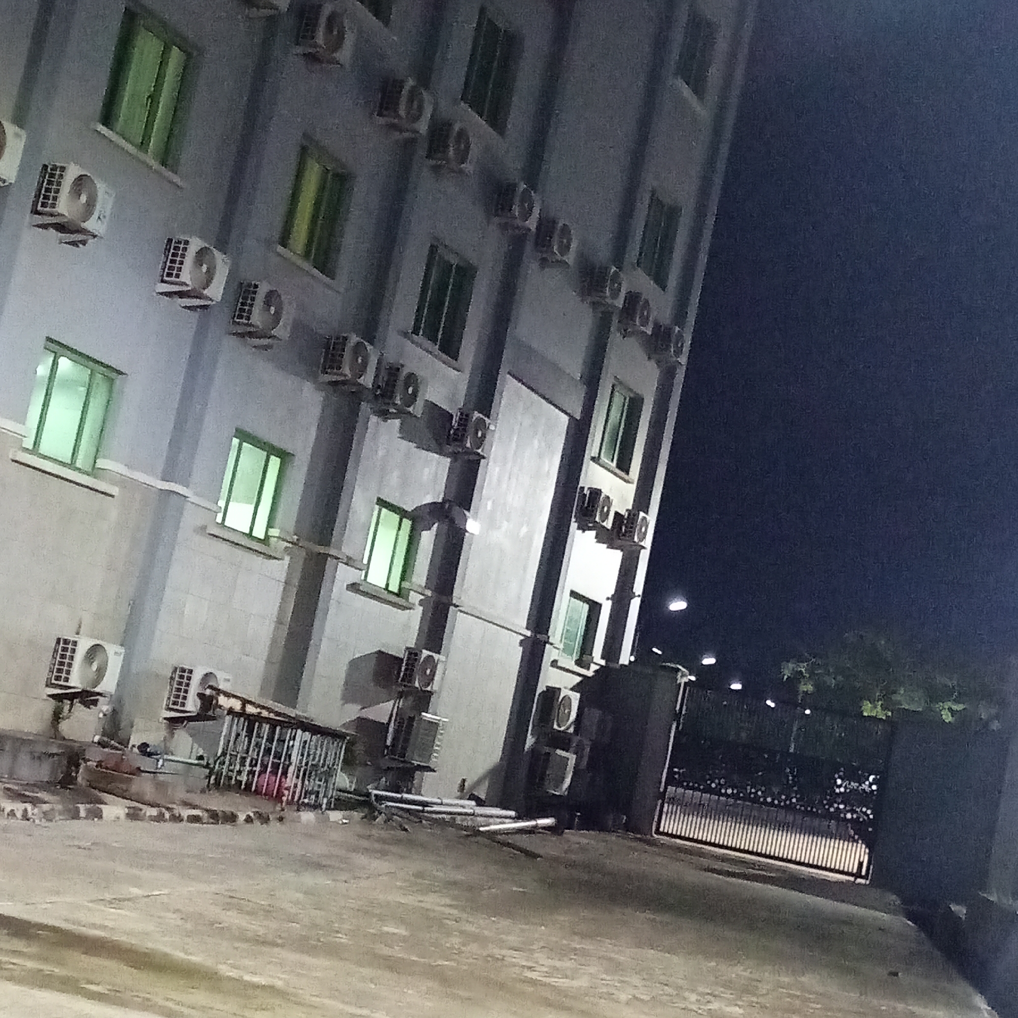 The image depicts the exterior of a multi-story building at night in Ibadan, Nigeria. The building has a gray facade and is adorned with numerous air conditioning units mounted on its exterior walls. Several windows with green-tinted glass are visible. The ground in front of the building is paved and appears to be a driveway or courtyard. A metal gate is visible on the right side of the image, with lights illuminating the area. The scene suggests a commercial or residential building, possibly an office or apartment complex. There are no people visible in the image. The overall atmosphere is dimly lit due to the nighttime setting, with artificial lighting from the building and streetlights creating highlights and shadows.