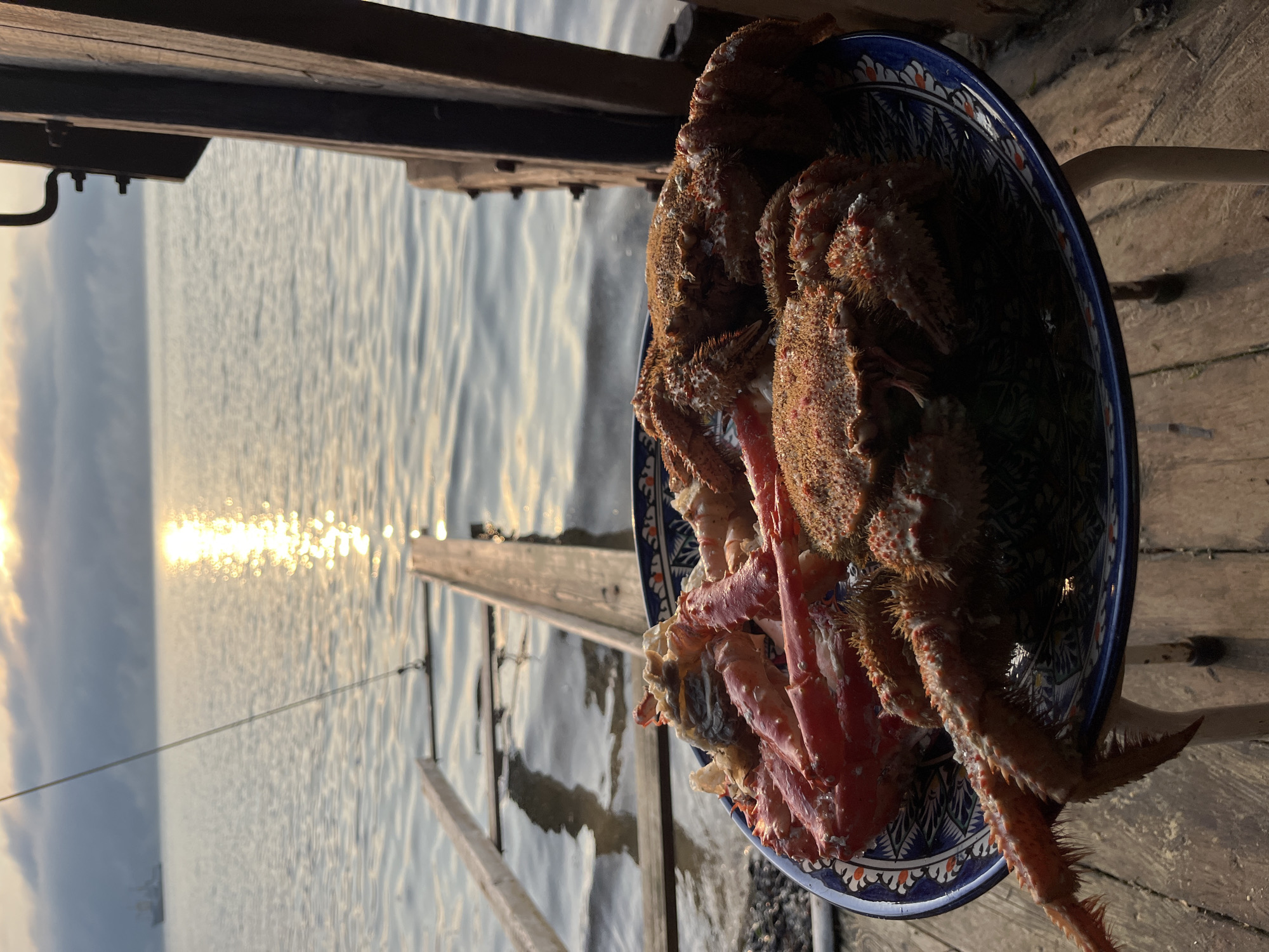 The image shows a plate of cooked crabs on a wooden surface by the water. The crabs appear to be a mix of red king crab legs and hairy crabs. In the background, the sun is setting or rising over the water, casting a golden reflection. There is a wooden ramp or ladder leading into the water, and a ship is visible in the distance. The scene is set at dusk or dawn. The location is identified as Sovetskaya Gavan', Russia, which is a port city on the Tatar Strait.