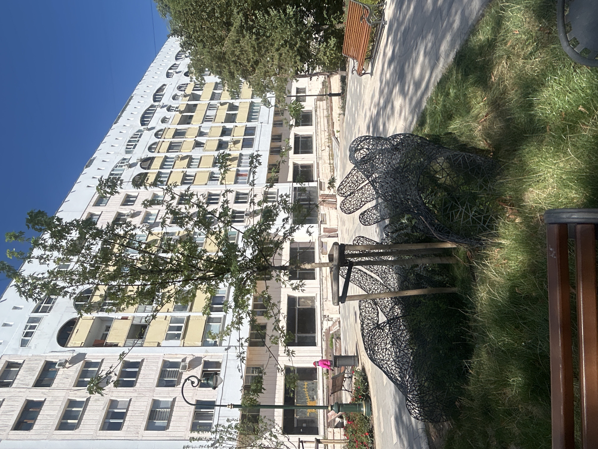 The image depicts an outdoor scene in the city of Almaty, Kazakhstan, characterized by a large, light-colored apartment building with yellow accents and numerous windows. In the foreground, a wire mesh sculpture resembling large hands rests amidst green grass. A young tree is planted to the right of the sculpture, and a park bench is visible further to the right, casting a shadow on the paved walkway. To the left of the sculpture, a lamppost with an ornate design stands next to a bench and a person wearing a pink jacket. The environment appears to be a public space, possibly a park or a courtyard, with a clear blue sky overhead.