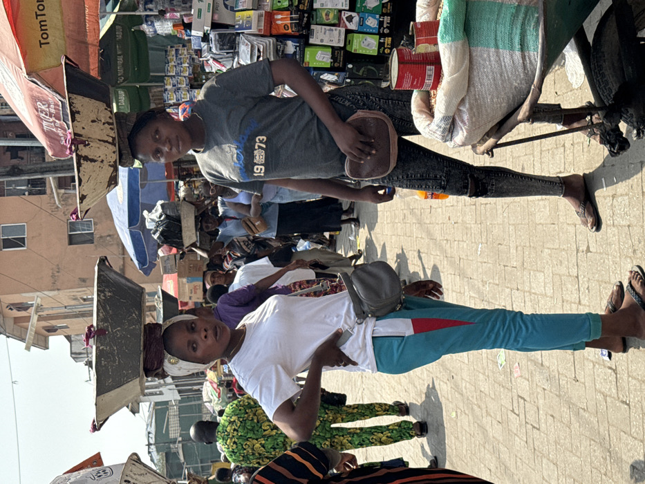 The image displays a bustling, crowded open-air market in Lagos, Nigeria, under clear daylight. Two women are central in the foreground. The woman on the left, with dark skin, wears a white short-sleeved t-shirt, teal and red-striped sweatpants, and black flip-flops. A gray fanny pack is slung over her shoulder, and a white headwrap with a gold embellishment secures a dark metal basin on her head, supported by a purple fabric ring. She stands with her right hand on her hip, looking slightly right. The woman on the right, also with dark skin, wears a dark gray short-sleeved t-shirt with "1973" text, distressed dark jeans, and dark flip-flops. A brown fanny pack is worn across her body, and a lighter, weathered metal basin rests on her head, supported by a dark fabric ring. She stands with her arms at her sides, looking slightly left.
Between and slightly behind the two women, a large white and green striped sack filled with grains, topped with red cans, is situated on a wheeled cart. An orange plastic bottle rests on the paved ground near the right woman's feet. The ground consists of light-colored interlocking bricks with minor scattered debris.
The background reveals numerous individuals navigating the crowded market. A man in a prominent green and yellow patterned garment is visible to the mid-left. Several people carry goods on their heads. Market stalls are shaded by red and blue umbrellas, one prominently displaying "TomTom" and "Tiger" branding. A stall to the right exhibits electronic accessories in clear packaging. Multi-story, light-brown buildings with visible windows form the distant background architecture.