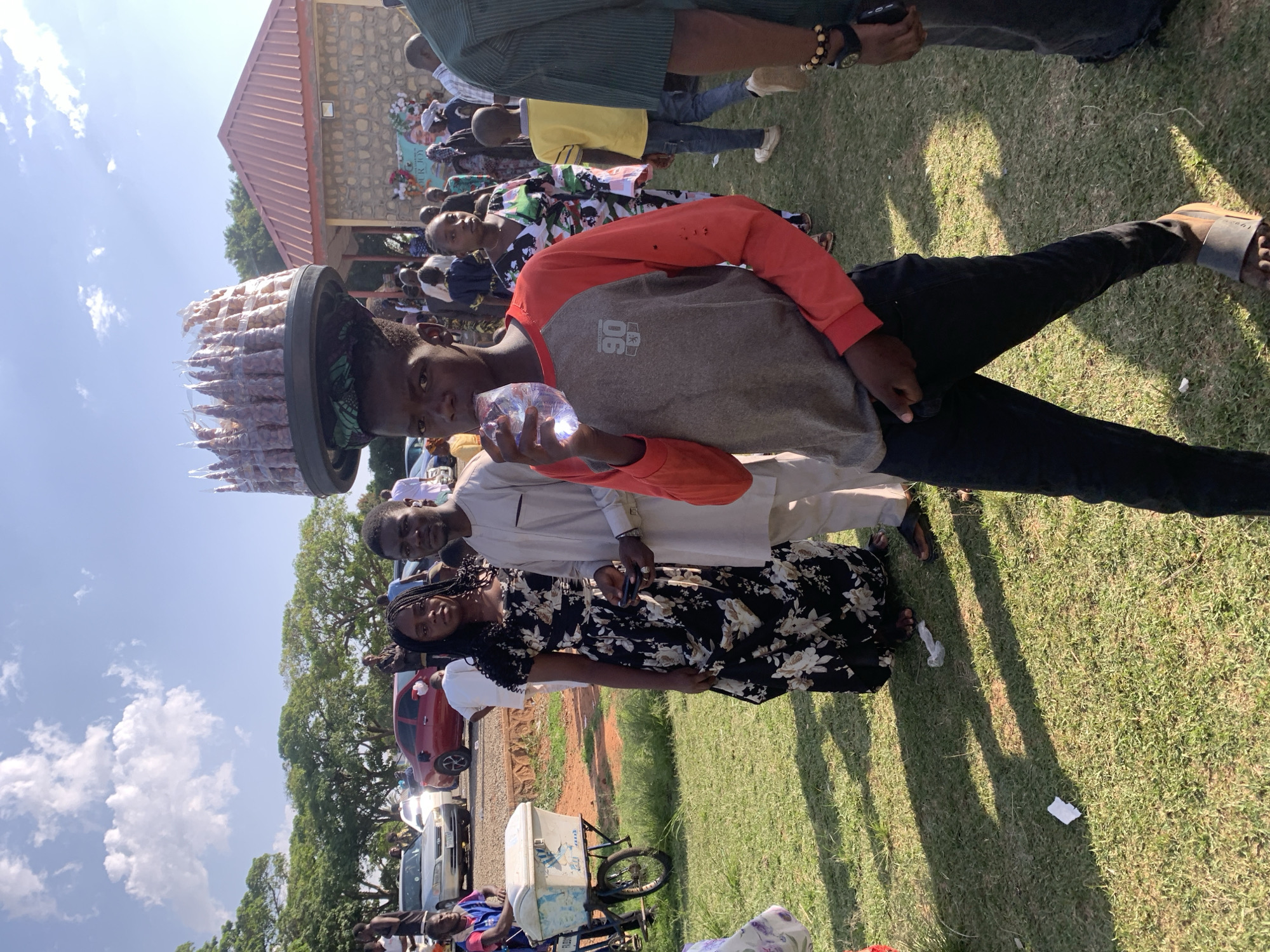 The image depicts a sunny outdoor scene in Jos, Nigeria, where a young man stands out with a unique display. He balances a large tray filled with bagged snacks or treats on his head while holding a clear plastic container, presumably water, in his hand. He is wearing a grey and red long-sleeved shirt, black pants, and sandals. Behind him, a diverse group of people are gathered, including a woman in a black and white floral dress and a man in a light-colored suit. In the background, there's a building with a red roof and a stone facade, along with parked cars and green grass, suggesting a public event or gathering. An ice cream vendor is also present.