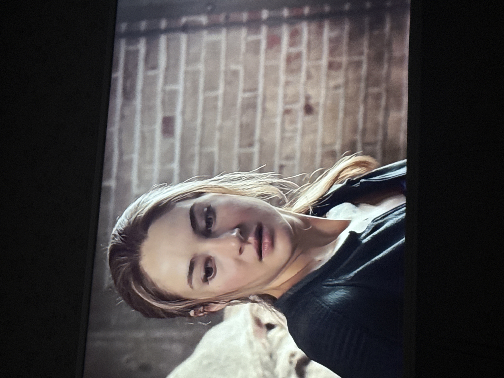 A medium shot shows a woman with fair skin and blonde hair, appearing somewhat disheveled. She wears a dark-colored top. Behind her, a brick wall is visible. The image appears to be a still from a film or television show. There is no evidence to suggest the image was taken in Mytishchi, Russia.