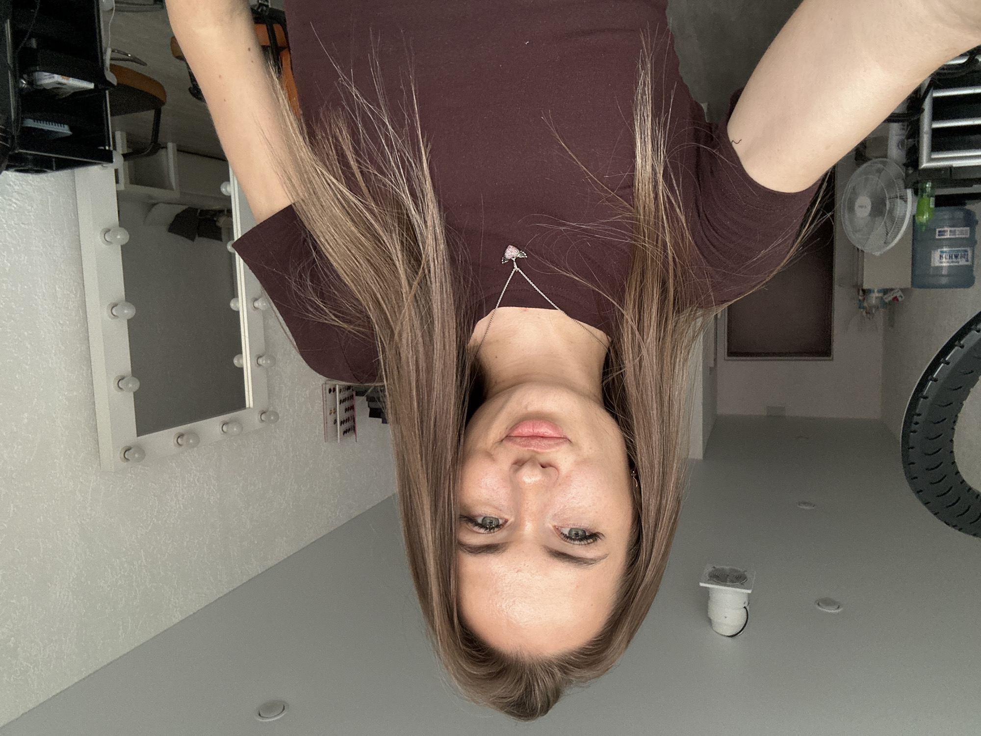 A young woman with long, straight, light brown hair is taking a selfie. She is wearing a dark brown t-shirt and a silver necklace with a heart-shaped pendant. She has light skin, green eyes, and is wearing makeup. She is indoors, in what appears to be a room with white textured walls. To her left, there is a large ring light, a water cooler, and a fan. To her right, there is a white mirror with lights around it, and a black trolley. The background shows a doorway and a couch in what looks like a waiting area. The scene is lit by artificial lighting.