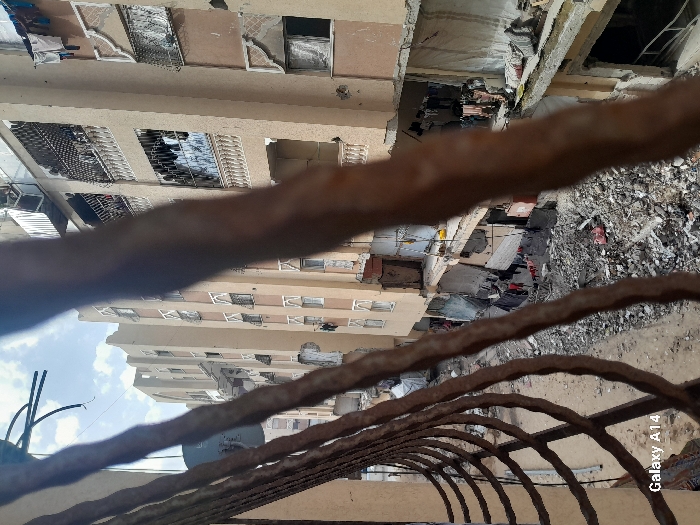 The image presents an elevated, oblique view of a severely damaged urban residential area in Khan Yunis, Palestinian Territory. The immediate foreground is partially obstructed by several out-of-focus, rusty, and twisted metal bars, consistent with a damaged balcony railing.
In the midground and background, multiple multi-story buildings, primarily constructed of light beige or tan concrete, line an open space. These buildings exhibit extensive structural damage: numerous windows are broken, missing, or covered with makeshift materials; facades display cracks and crumbling sections; and several balconies show signs of collapse or missing railings. On an upper-level balcony of one building, approximately 5-7 individuals, dressed in light-colored clothing, are visible. Clothes are seen hanging to dry from various balconies and ground-level structures.
The ground area between the buildings is covered in a significant amount of rubble, shattered concrete, and debris, indicative of widespread destruction. Amidst the debris, remnants of smaller structures, tarpaulin coverings, and scattered personal belongings are visible. The sky above is a light blue with scattered white clouds. The scene depicts a post-destruction urban environment with visible signs of ongoing human habitation amidst the damage.