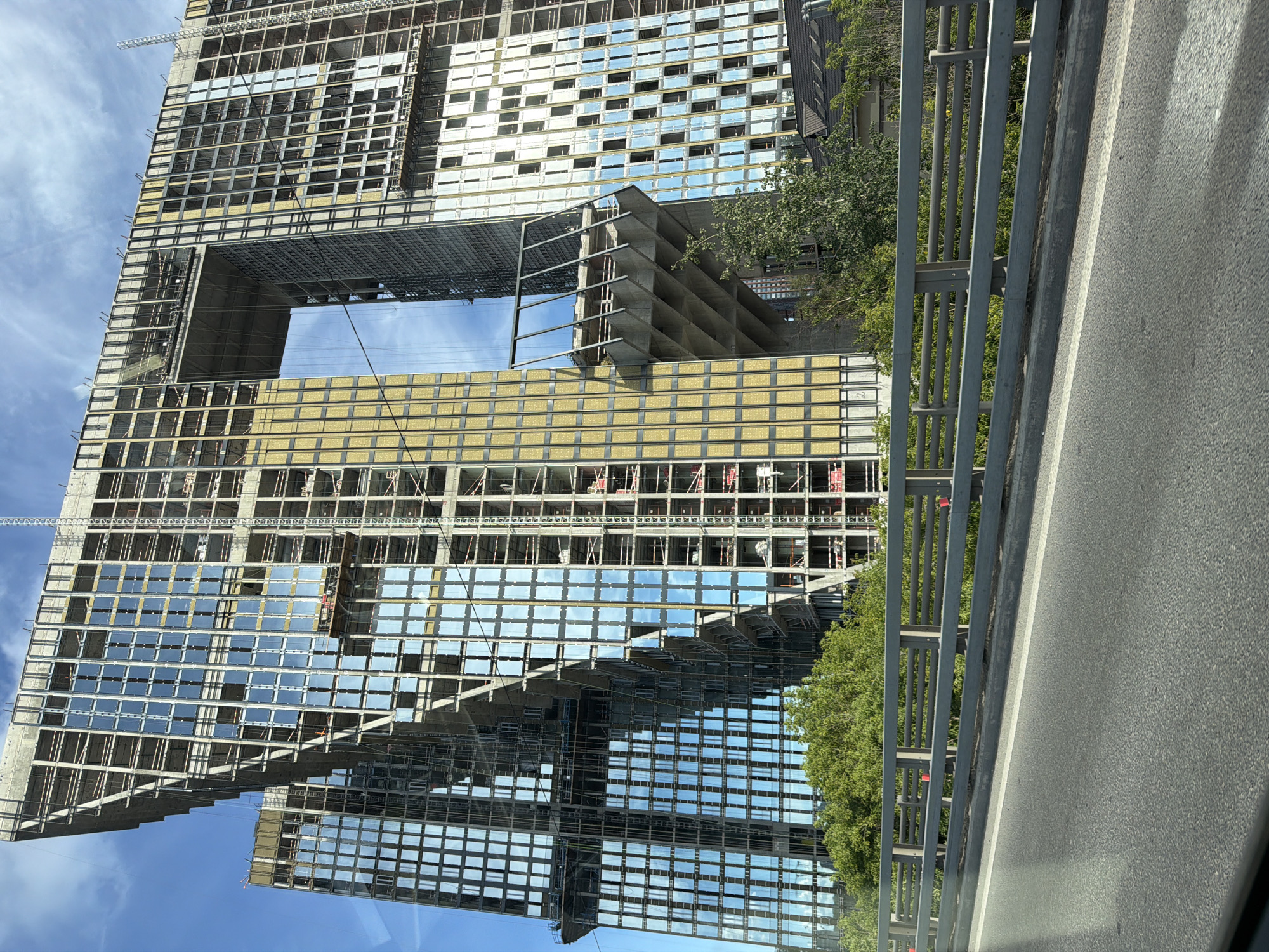 The image shows a large, modern building under construction in Khimki, Russia. The building has a unique architectural design with two main towers connected by a large open space at the top. The facade is largely glass, reflecting the blue sky and clouds. Construction scaffolding is visible on the exterior, and workers can be seen on platforms. The building is surrounded by lush green trees. In the foreground, there is a road with a grey metal railing, suggesting the photo was taken from a vehicle or a pedestrian bridge. The overall impression is one of significant urban development and ongoing construction.