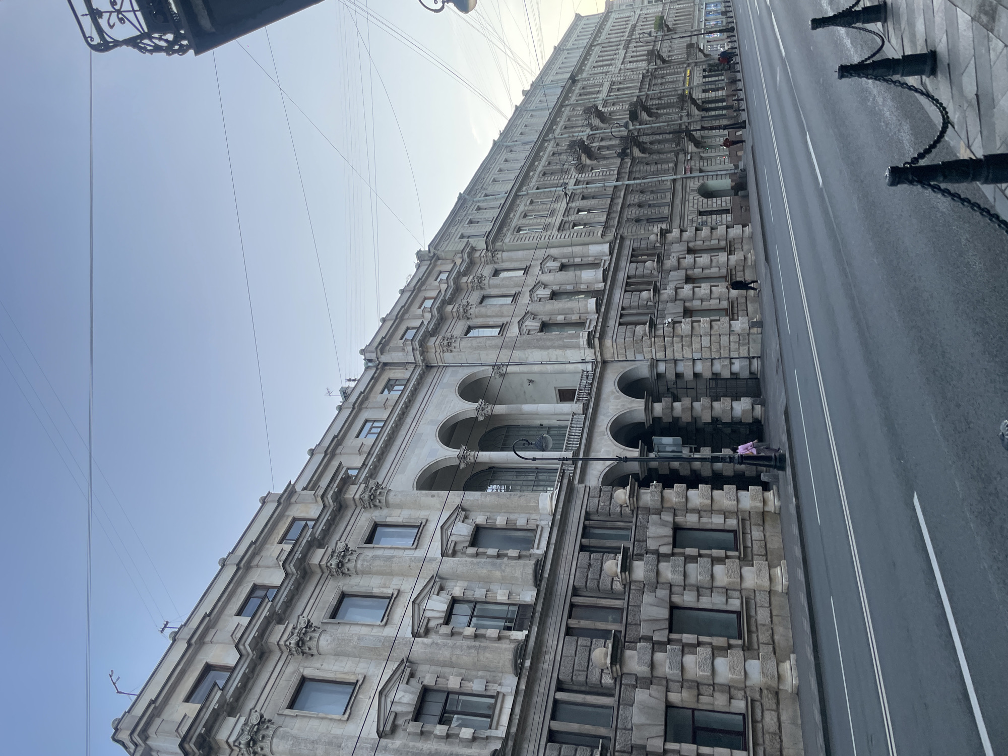 This image depicts a wide street in Saint Petersburg, Russia, with a long, multi-story building lining its left side. The building exhibits classical architecture with ornate detailing, including columns, arches, and decorative window frames. The street itself is paved and appears to be an active thoroughfare, with lane markings visible. A few scattered people are present on the sidewalk in front of the building, some walking and one person in a pink shirt standing near a lamppost. The sky is clear and overcast, suggesting either early morning or late evening light. The overall scene conveys a sense of urban environment with a historical architectural aesthetic.