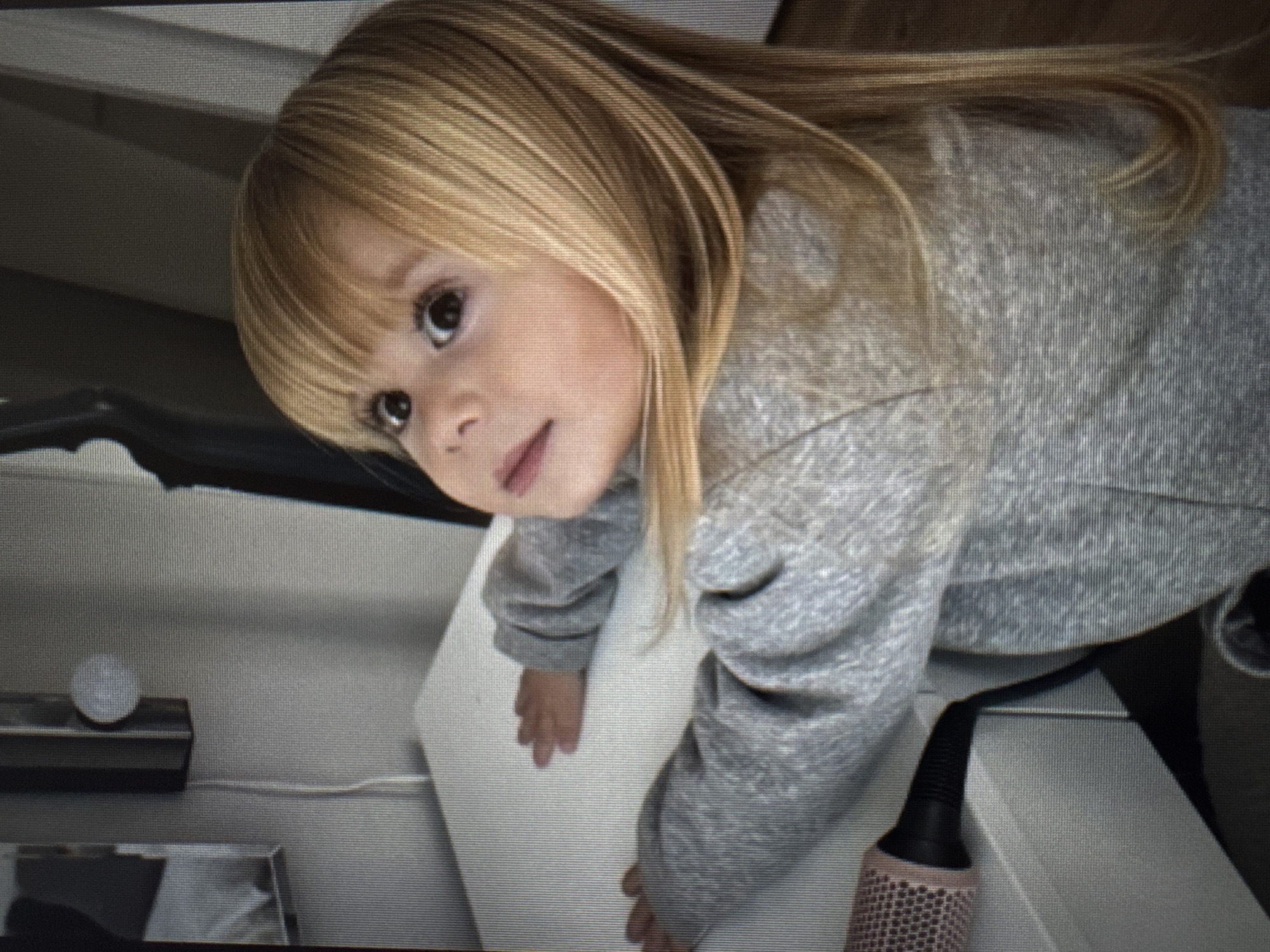 A young child, appearing to be a toddler or preschooler, with long, straight light blonde hair and bangs, is the central subject of the image. She has wide, dark eyes and a neutral expression with slightly parted lips. The child is wearing a light gray, possibly heathered, long-sleeved sweatshirt. She is leaning forward and looking upwards, with her hands placed flat on a white, soft surface, possibly a cushion or mattress.
The activity suggests a moment of curiosity or playful interaction as the child looks directly into the camera.
Notable details include an indoor setting, likely a home, with hints of furniture such as a dark headboard or similar structure in the background. To the left of the child, a black rectangular object with a small white circular item on top is visible, along with a white cable extending from it. To the right, partially visible, is a pink and black cylindrical object, resembling a hairbrush or hair roller. The overall image has a noticeable screen-door effect, with horizontal lines and pixelation, indicating it is likely a photograph taken of a digital screen. There is no visible text.
