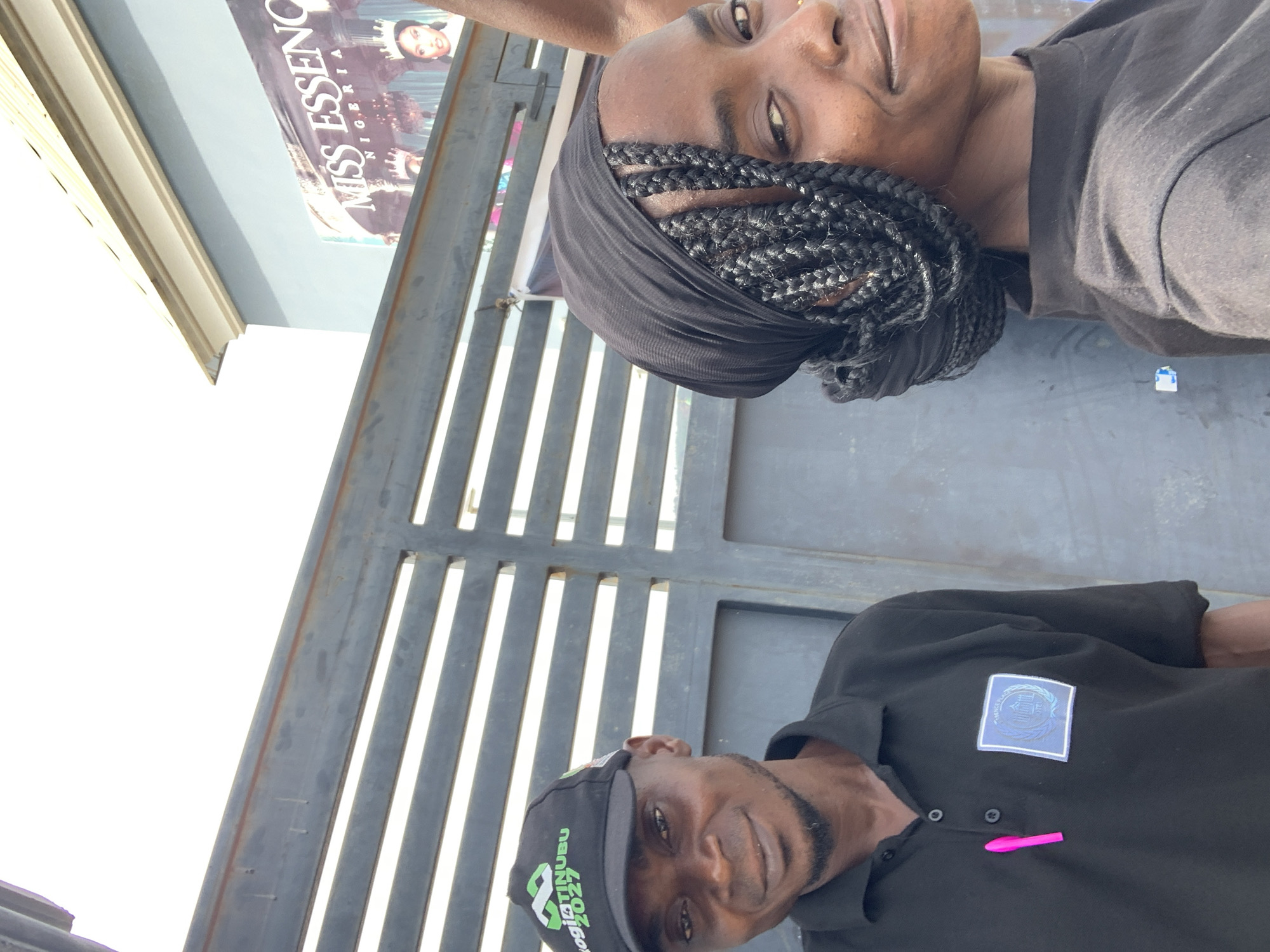 The image depicts two individuals outdoors. A woman with braided hair wrapped in a black head covering is positioned at the top of the frame. A man in a black polo shirt and a black cap with green and white lettering is at the bottom. A pink object resembling a pen is visible on the man's shirt. Behind them is a metal gate with horizontal bars. A poster is visible in the background, with a title that includes "MISS ESSENCE NIGERIA". Given the available information, the location is likely Adankolo, Nigeria.