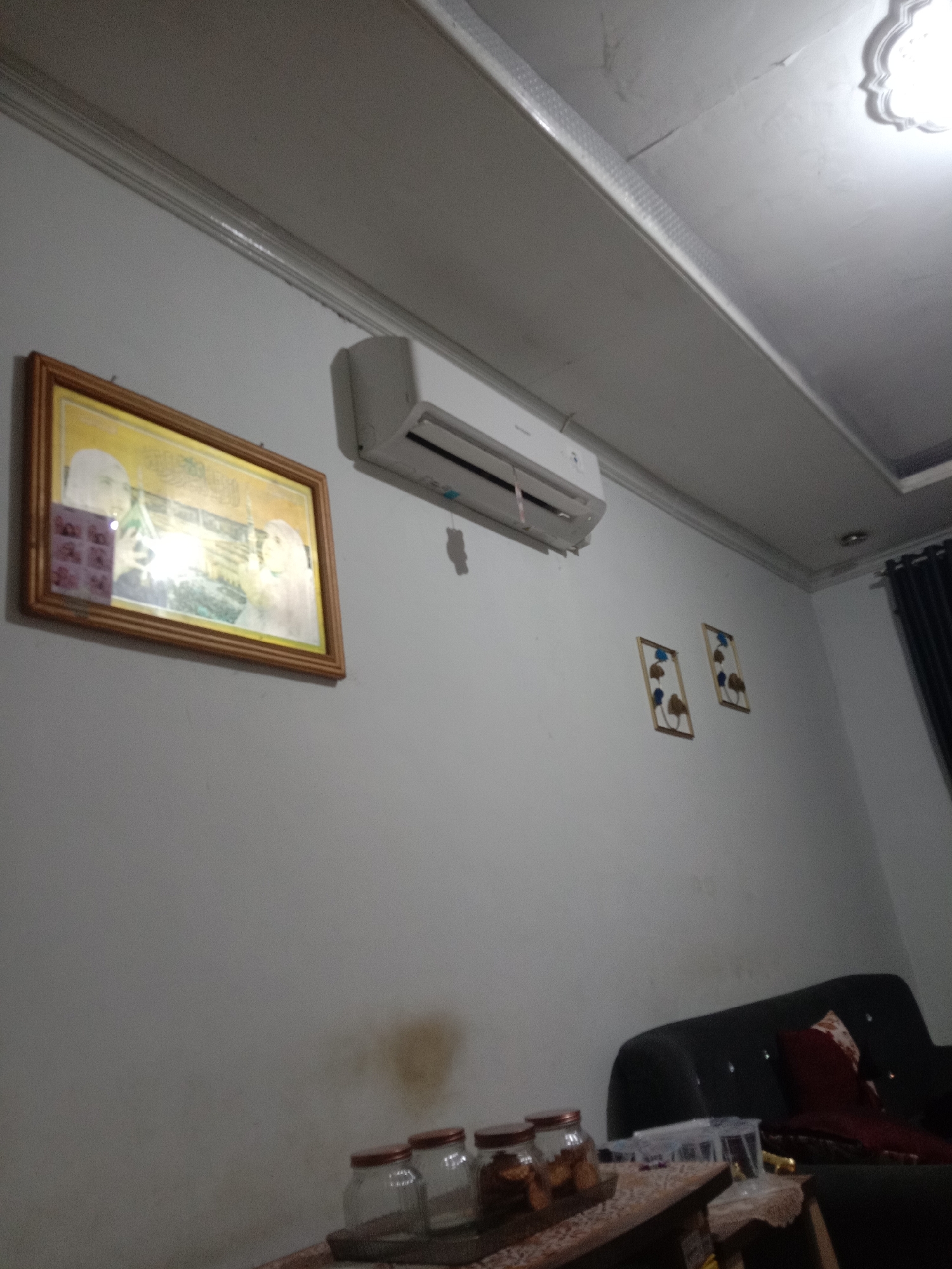 The image shows a sparsely decorated indoor scene, likely a living room. A white air conditioning unit is mounted on a light-colored wall. To the left of the air conditioner, a framed picture with a gold border hangs on the wall, depicting what appears to be a religious scene or landmark. Below the air conditioner, two small framed decorative items with abstract designs are also affixed to the wall. In the lower right portion of the image, a dark-colored sofa with a red cushion is visible. In the foreground, a wooden surface holds several glass jars with red lids, some containing what looks like food items or snacks. The overall lighting suggests it is daytime, with a ceiling light fixture visible in the upper right corner. The setting is consistent with a residential interior in Kwandang, Indonesia.