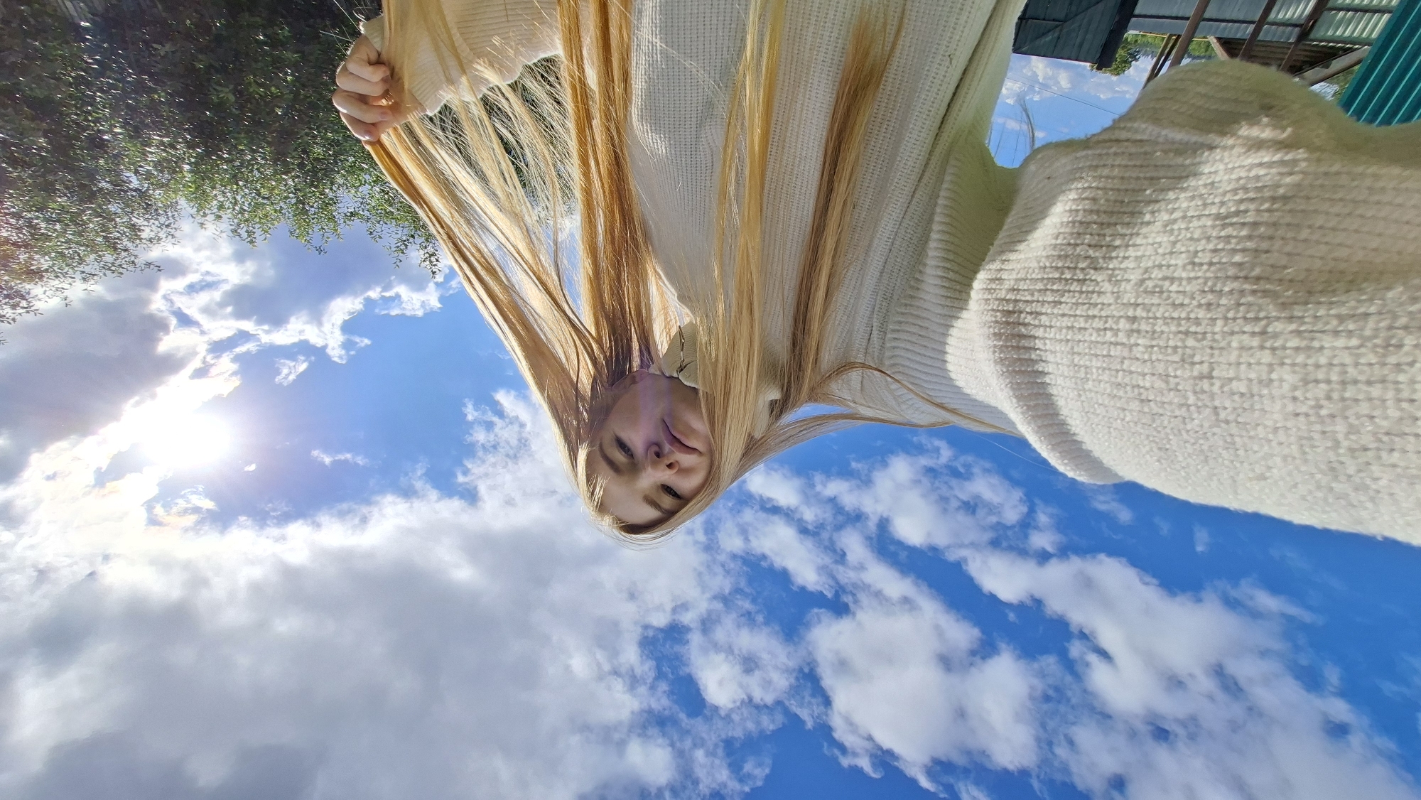 This is a close-up, upside-down selfie taken from a low angle, looking up at a bright blue sky filled with fluffy white clouds. The sun is shining brightly, creating lens flare in the upper left portion of the image.
The main subject is a young woman with long, blonde hair, wearing a cream-colored, textured sweater. She is holding a lock of her hair in her left hand, pulling it upwards. Her face is turned towards the camera, with her eyes looking slightly upwards and a relaxed expression.
In the background, above the clouds, some greenery is visible on the left, suggesting trees. On the right, partially visible at the top, are parts of a building with a corrugated metal roof and a green structure. These elements, along with the general brightness of the scene, suggest a daytime setting. The weather appears to be clear and sunny with some clouds.
The upside-down perspective, combined with the upward gaze into the sky, creates a playful and perhaps carefree feeling. The location context provided (Большой Букор, Russia) hints at an outdoor setting, possibly a yard or open area near a dwelling. There is no visible text in the image.
